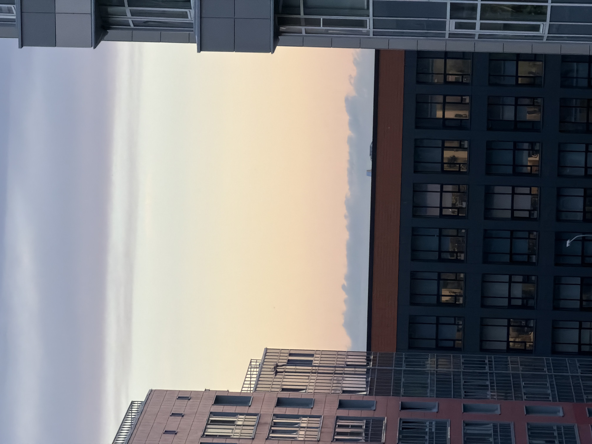 The image captures a view of modern buildings against a gradient sky, transitioning from pale blue at the top to soft pink and yellow near the horizon, suggesting either dawn or dusk. On the left, a tall, light pink building with numerous windows and a decorative railing on its upper level is visible. Adjacent to it, and slightly set back, is a darker, rectangular building with a row of orange-brown material along its top edge and a facade of large, dark-framed windows, some of which show interior lights. To the right, a portion of a grey building with a sleek, contemporary design and large glass windows is also present. The sky above the buildings is mostly clear, with thin, wispy clouds near the horizon, also reflecting the soft hues of the sky. The overall scene depicts an urban landscape with contemporary architecture under a twilight sky.