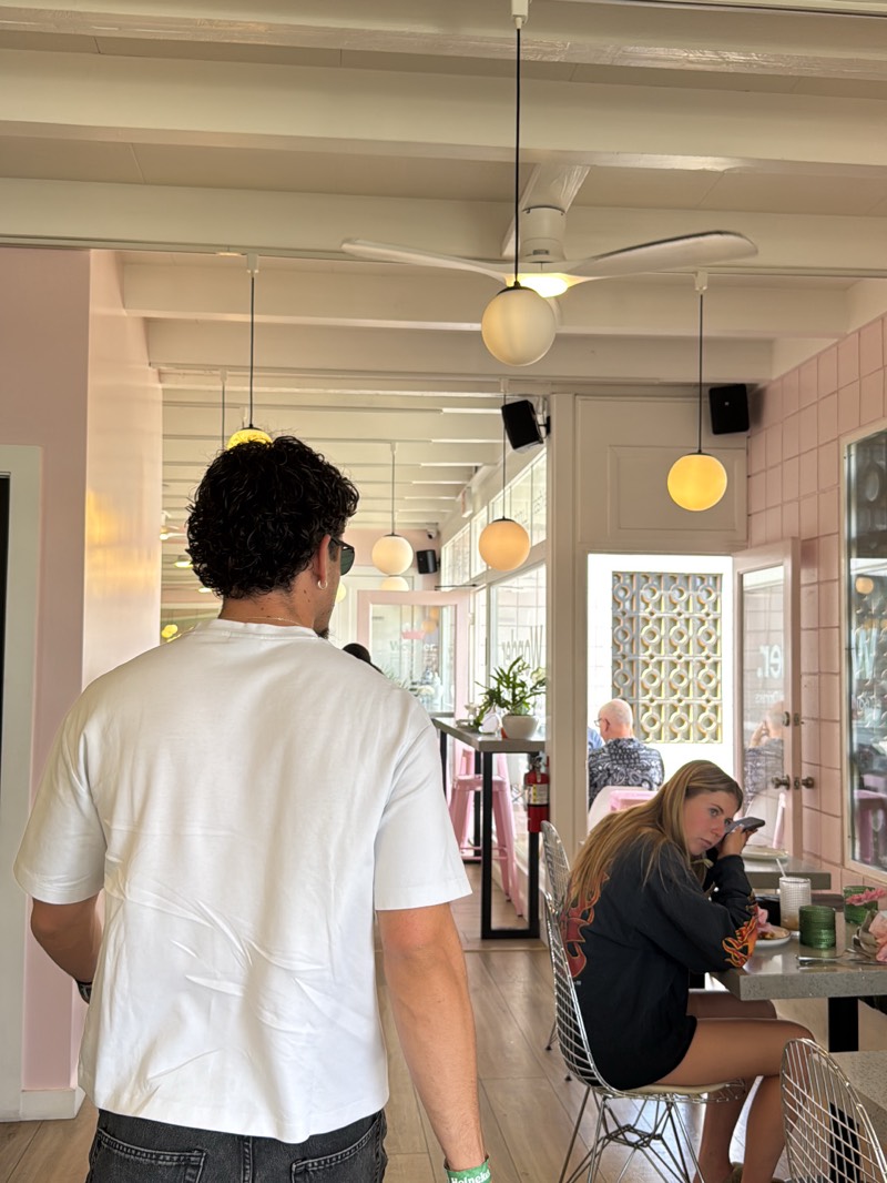 The photograph, taken indoors, shows a man with dark curly hair and sunglasses walking away from the camera. He is wearing a white t-shirt and dark jeans. In the background, a young woman with blonde hair is seated at a table, looking towards the camera while holding a phone to her ear. She wears a dark shirt with a flame design. The setting appears to be a restaurant with pink tiled walls and white ceiling beams. A ceiling fan with a spherical light fixture is visible. Other patrons can be seen in the background. The restaurant setting and the presence of a Heineken bracelet on the man's wrist suggest a casual atmosphere. The location is likely in Palm Desert, United States.