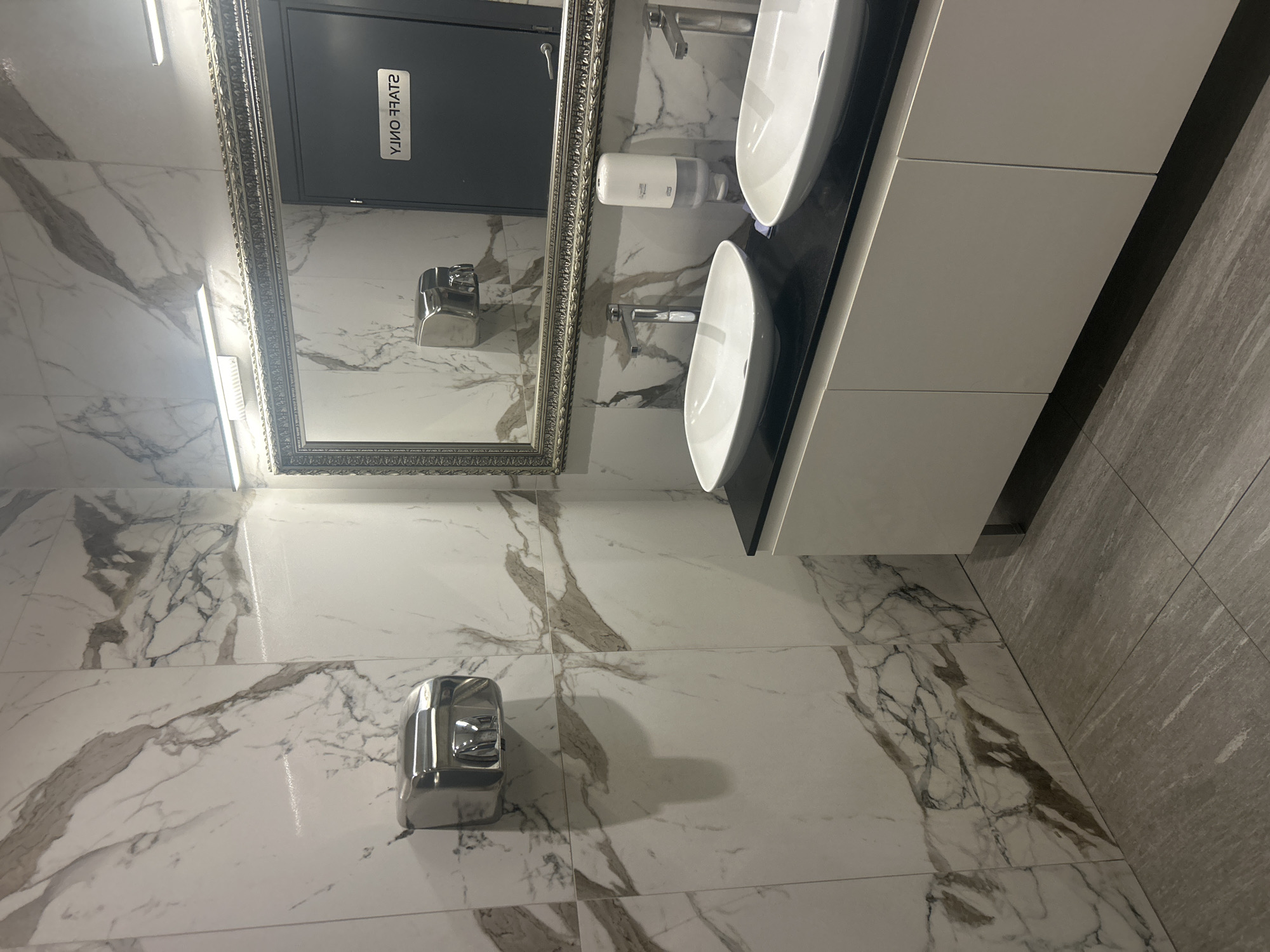 The image shows a modern bathroom with white marble-patterned tiles on the walls and floor. Two white oval sinks are mounted on a dark countertop, with chrome faucets. A white automatic soap dispenser is positioned between the sinks. To the left, a chrome hand dryer is mounted on the tiled wall. In the background, a large, ornate mirror reflects a gray door with a sign that reads "STAFF ONLY" in white lettering. The lighting appears to be artificial, likely overhead lights or integrated into the mirror. The overall scene suggests a clean and well-maintained public restroom, possibly in a commercial establishment. There are no people visible in the image. The time of day and weather are not discernible. The location context is Plovdiv, Bulgaria.