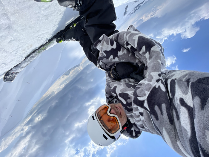 An inverted image depicts a single individual on a snow-covered mountain slope at Katun Bačko brdo, Montenegro. The subject is positioned upside down, with their head in the lower frame and feet with skis in the upper frame. They are wearing a white ski helmet with amber-tinted goggles, a grey, black, and white camouflage-patterned fleece jacket, black ski pants, and black ski boots. Black skis with green accents are partially visible, secured in the snow. The person is looking towards the camera with a slight smile. The background consists of a blue sky with scattered white clouds, and distant snow-covered mountain ranges featuring multiple small, dark, upright structures, consistent with ski lift pylons. The ground is uniformly covered in snow.