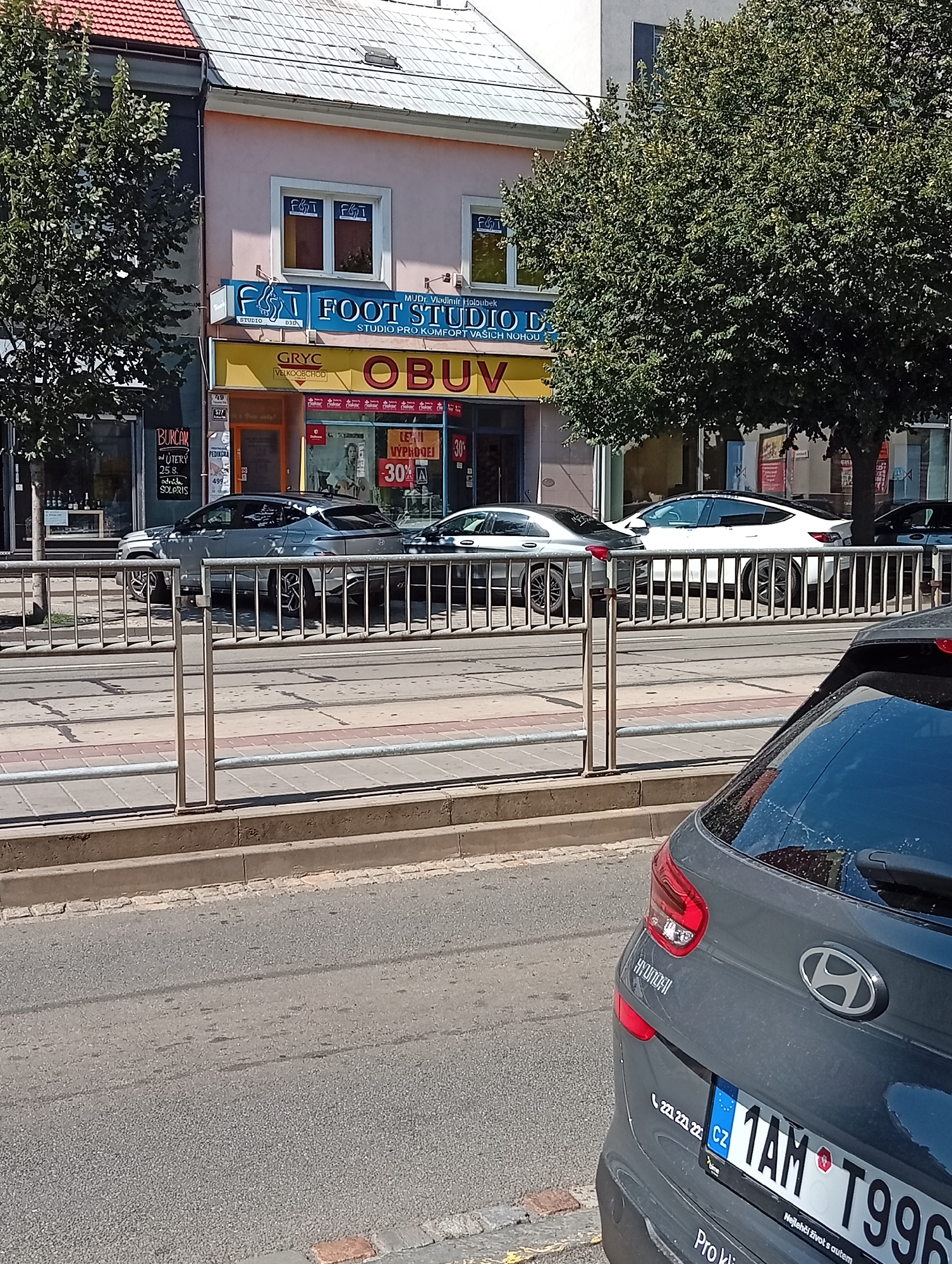 In Brno, Czechia, a street scene unfolds with a focus on a shop front and the passing traffic. The shop, named "FOOT STUDIO PRO KOMFORT VAŠICH NOHOU," is painted pink and features a yellow awning with the word "OBUV" (shoes in Czech). Above this, a blue sign displays "FOOT STUDIO." Adjacent to the shop are display windows showcasing various shoe brands.
Several vehicles are visible on the street. A dark grey Hyundai SUV is parked in the foreground, its rear end prominent in the shot. Further down the street, a white Tesla Model 3 is parked, its sleek design evident. To the left of the Tesla, a silver sedan, possibly a BMW, is also parked. The vehicles are positioned on a road that appears to be a streetcar line, indicated by the tracks in the foreground.
The environment is urban, with buildings lining the street. Trees with lush green foliage are present, partially obscuring the upper floors of the buildings. A metal railing separates the sidewalk from the road. The weather appears to be sunny, casting distinct shadows on the pavement. The overall impression is a typical street scene in a European city, likely during daytime.