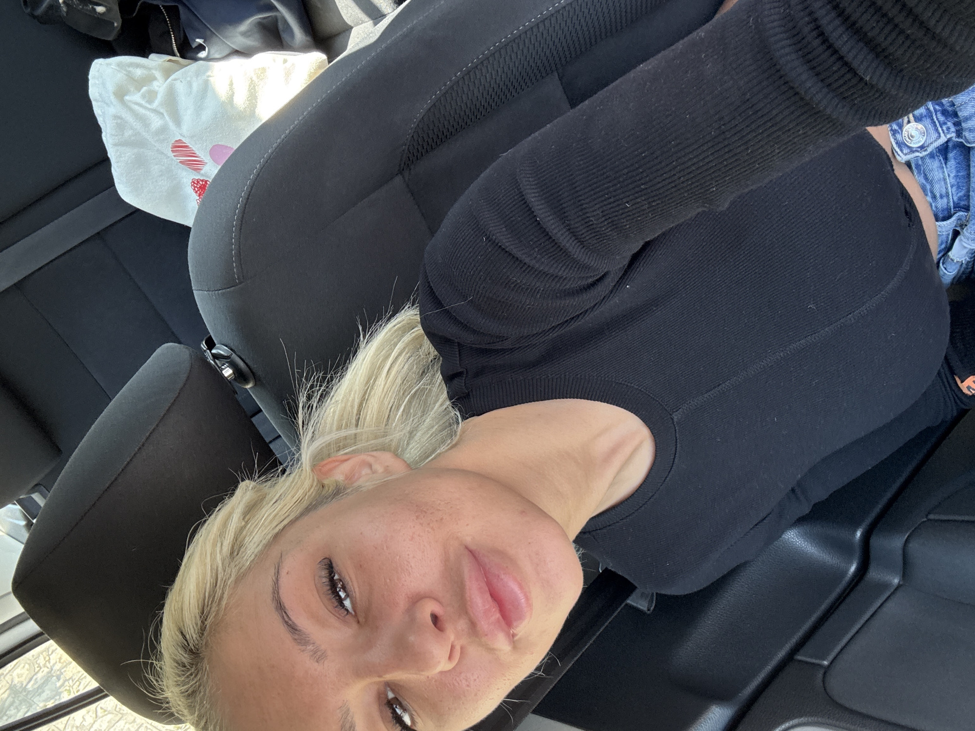 A selfie taken inside a car. A woman with blonde hair and a black long-sleeved shirt is visible. The car seat and interior are also visible. A white tote bag with a pink and red design is on the seat beside her. The location is not discernible and cannot be determined to be in Plovdiv, Bulgaria.