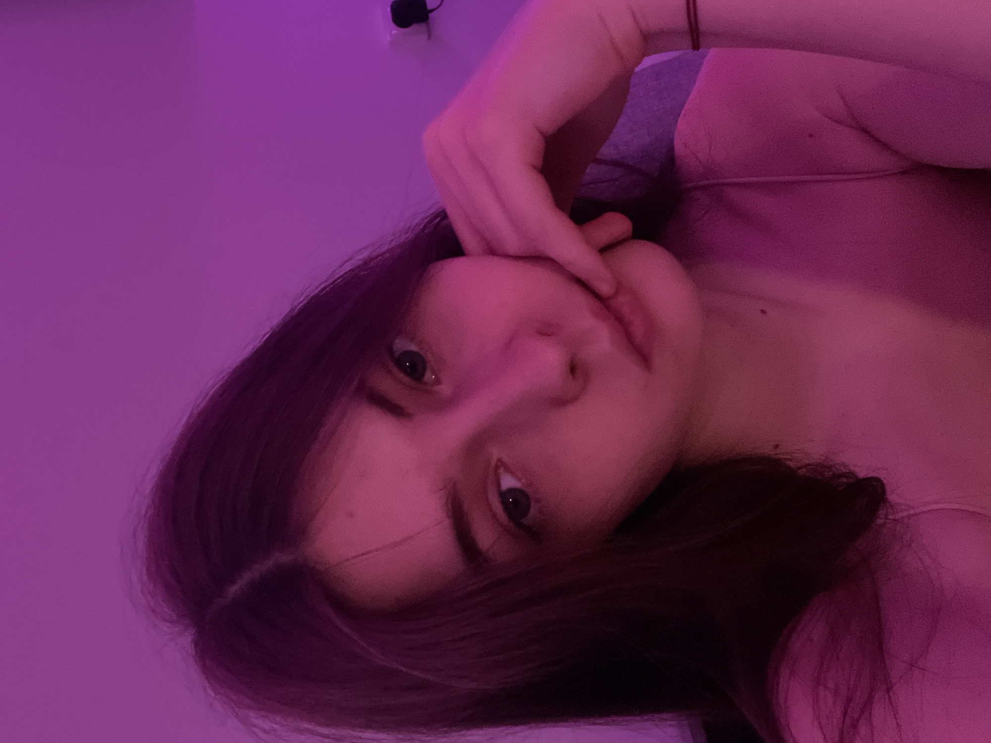 A young woman with dark hair is visible from the shoulders up, appearing to pose for a selfie in a room illuminated by purple light. She rests her hand near her face, and the angle of the image suggests it was taken from a slightly elevated perspective. A dark object is visible on the wall behind her. There is no information available about the location being Saint Petersburg, Russia.
