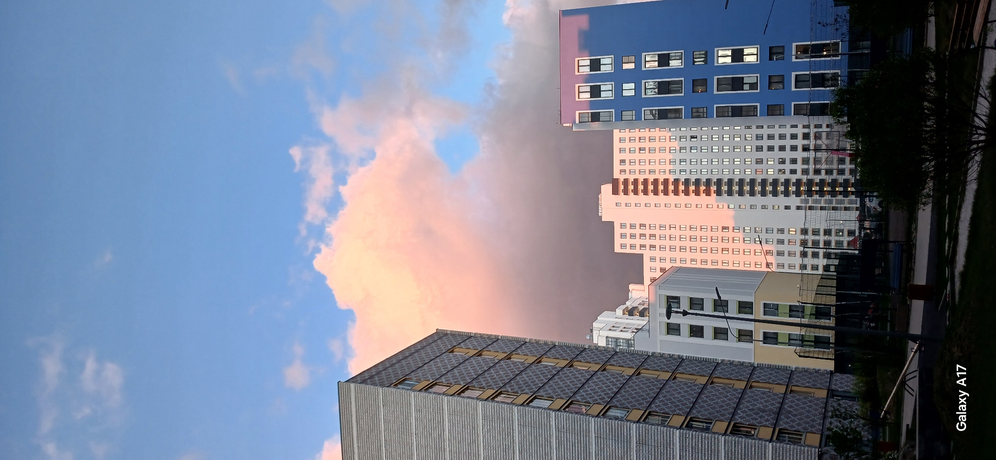 The image depicts an urban landscape in Yekaterinburg, Russia, with a focus on modern high-rise buildings against a backdrop of a dramatic sky. The buildings are of varied architectural styles, including structures with blue facades, white facades, and gray facades with patterned detailing. The sky is dominated by pink and gray clouds illuminated by the setting sun, contrasting against the clear blue sky. The image is taken from a low angle, emphasizing the height of the buildings. Vegetation and some street furniture like lampposts are partially visible, suggesting a park or residential area. The text "Galaxy A17" is visible on the lower right corner of the image.