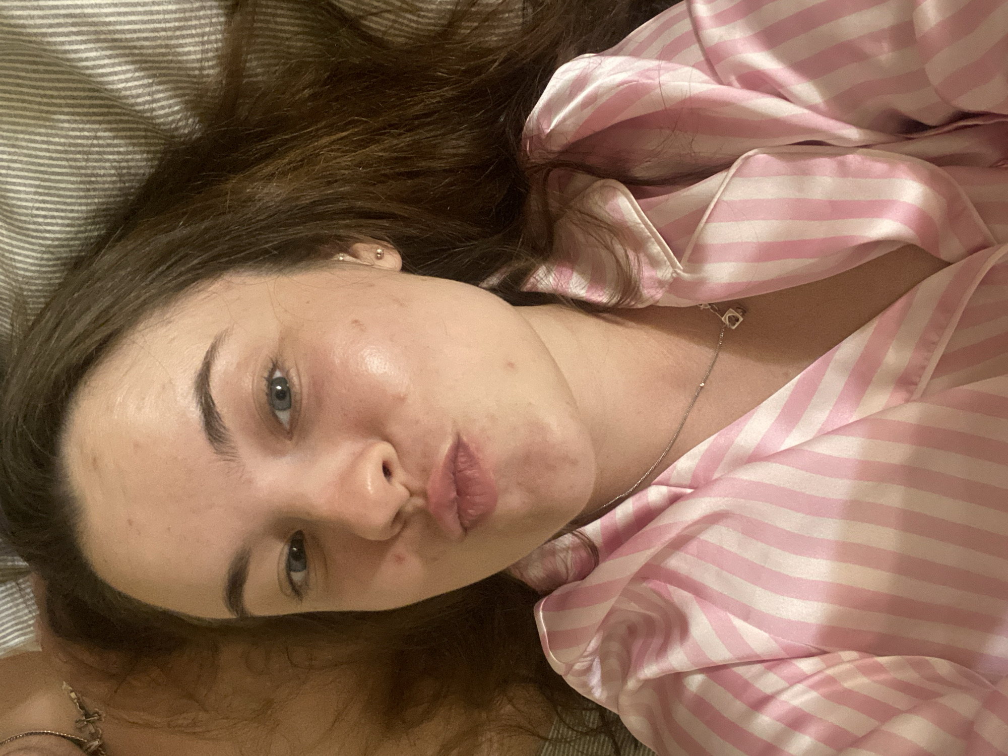 A young woman with long brown hair lies in bed, wearing pink and white striped pajamas. She is looking at the camera with her lips pursed, giving a kiss. A silver necklace with a small pendant hangs around her neck. The background is a striped pillowcase. The image was taken in Poltava, Ukraine.