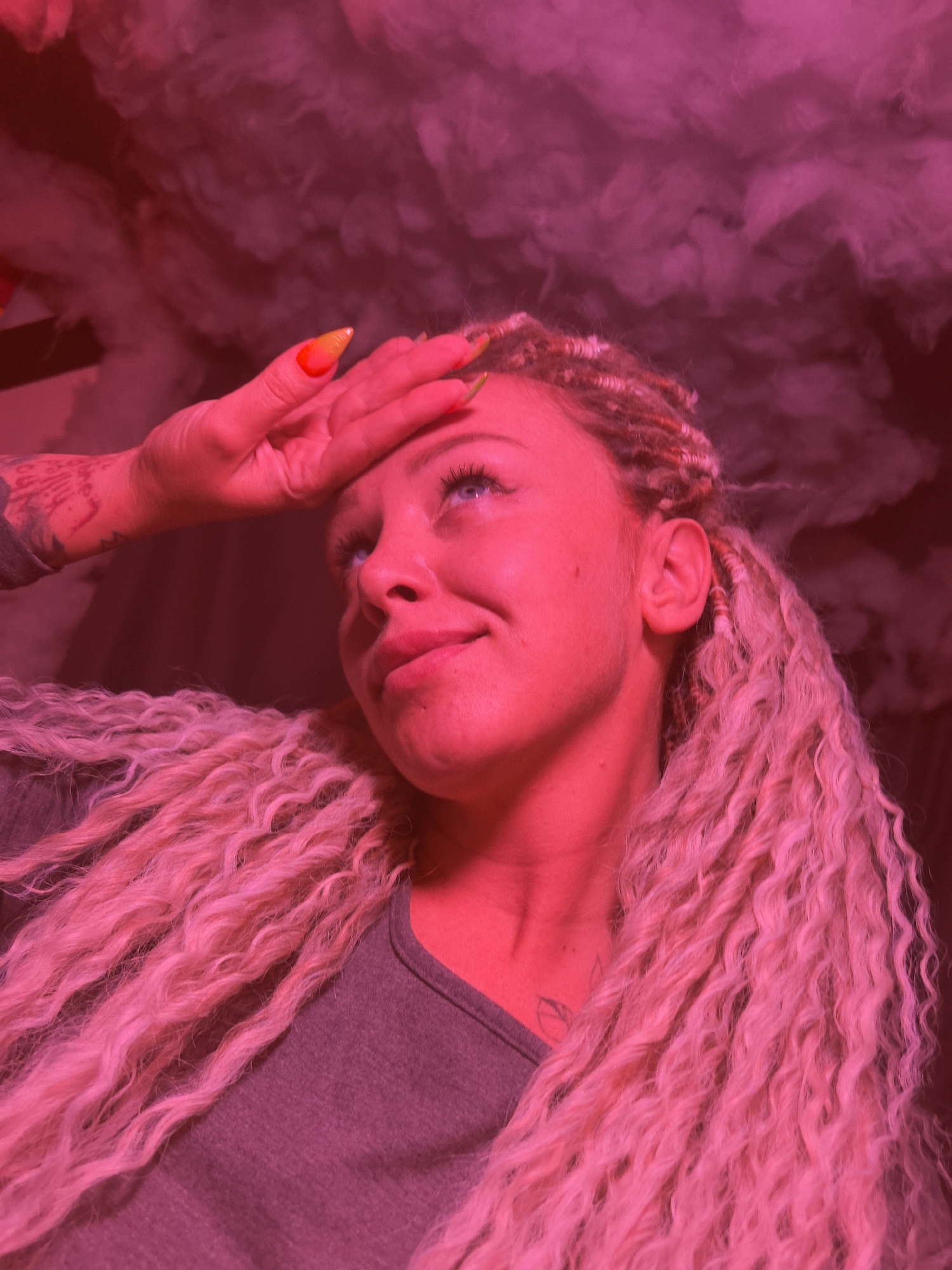 The media file displays a close-up portrait of a person, likely a woman, under dramatic red and magenta lighting. She has long, voluminous, light-colored curly or wavy hair, possibly mixed with dreadlocks or braids. Her blue eyes are visible as she looks upwards and slightly to the right, her expression appearing thoughtful or slightly pensive, with a subtle smile. Her left hand is raised to her forehead, fingers spread, showcasing brightly colored, pointed nails with a yellow and orange gradient. A tattoo is visible on her left forearm. She is wearing a dark gray or charcoal-colored top. The background is dominated by a smoky or cloudy texture, heavily illuminated by the same red/magenta light, suggesting an indoor setting with artificial effects. There is no visible text. The intense lighting and the smoky atmosphere create a surreal or club-like ambiance. The image provides no specific visual cues to indicate the location as Noviy, Russia.
