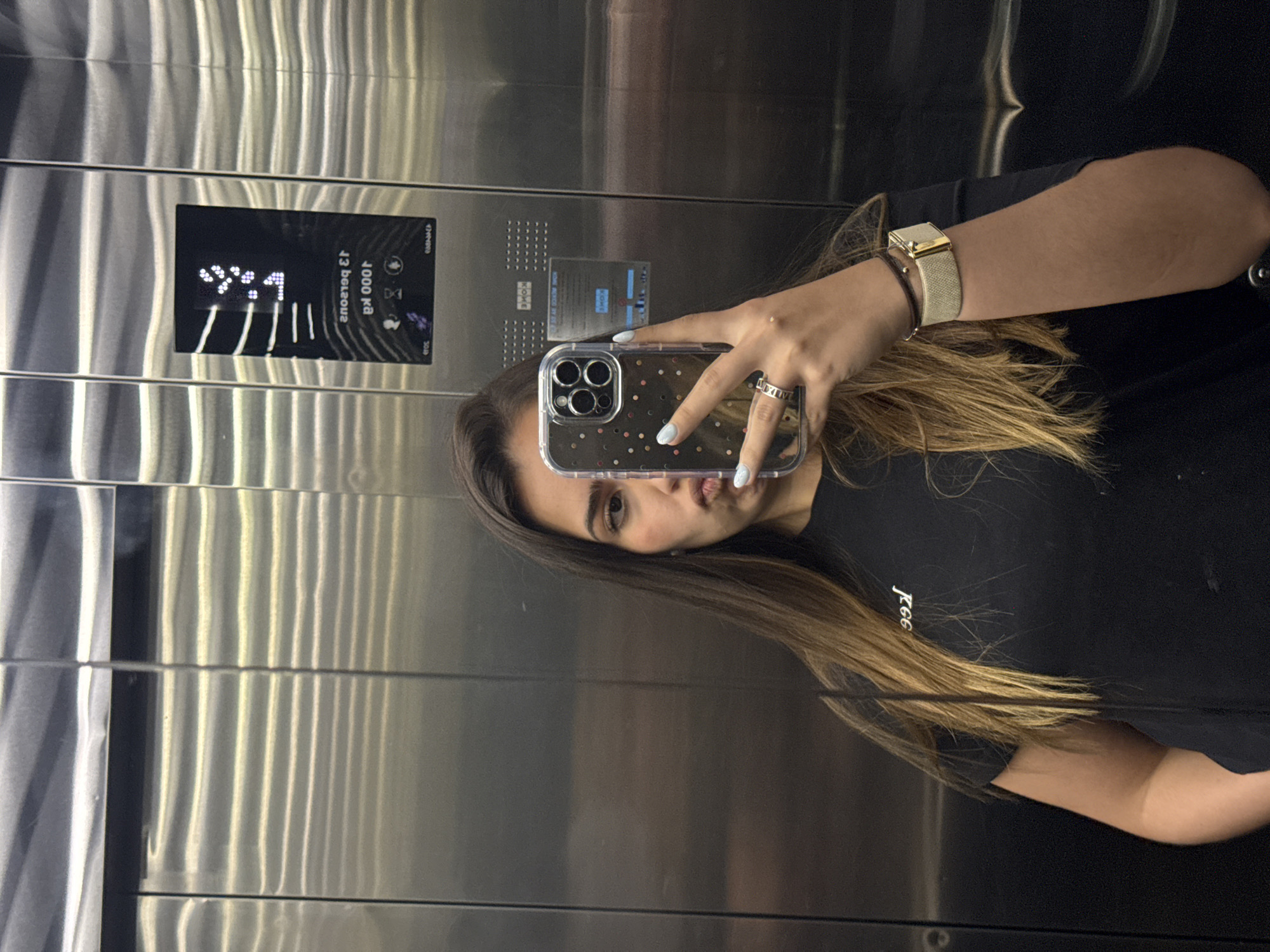 A woman is taking a selfie in an elevator. The elevator's panel indicates it can hold 1000 kg and is currently on the 13th floor. The woman has long brown hair, is wearing a black shirt and a watch, and holding a cell phone with a clear case. The elevator is metallic. The location is not specified and cannot be determined from the image.