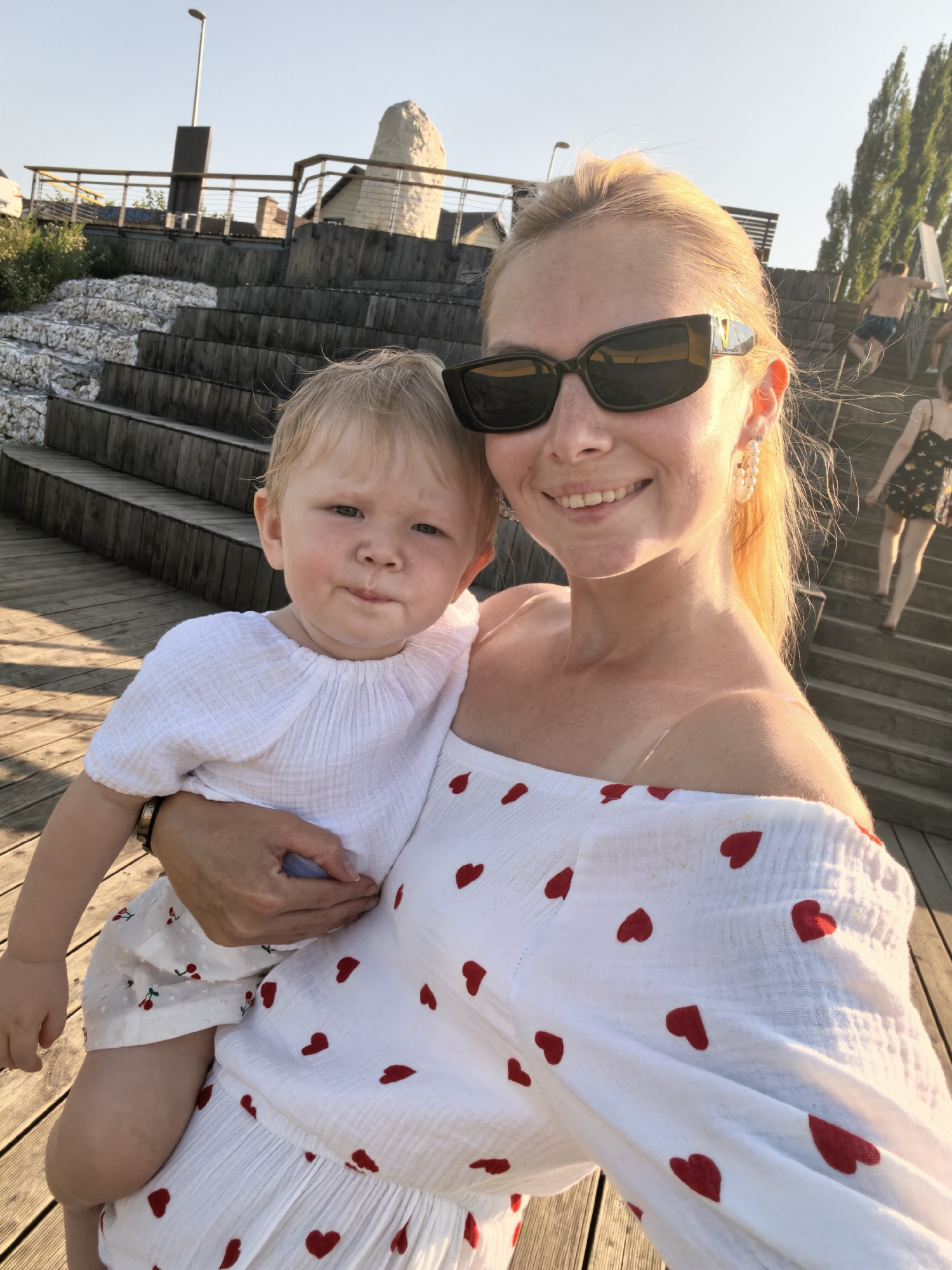 A woman wearing sunglasses and a white off-the-shoulder dress with red hearts is holding a baby wearing a white crinkled fabric outfit. The woman is smiling and looking at the camera, while the baby looks slightly to the side with a puckered expression. They are standing on a wooden boardwalk, with wooden tiered steps and stone-filled retaining walls behind them. In the background, people are visible walking up the stairs. The scene appears to be outdoors in daylight, with a clear sky. The location is described as the city of Sterlitamak, Russia.