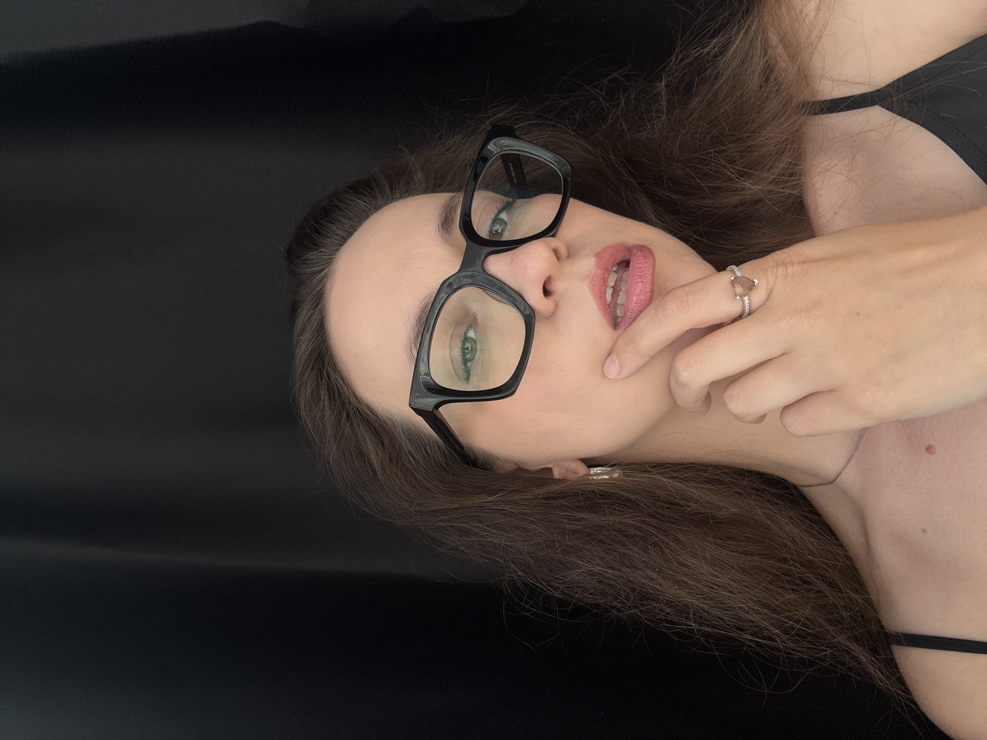 A young woman with long, dark brown hair and light skin is lying down against a black surface. She is wearing large, black-rimmed glasses and a black top. Her right hand is resting on her face, with her index finger touching her lips, which are painted with pink lipstick. A delicate ring with a pear-shaped stone and smaller stones adorns her ring finger. A fine silver necklace is visible around her neck. Her eyes, visible through her glasses, appear to be green. The overall mood is intimate and relaxed. No text is visible in the image.