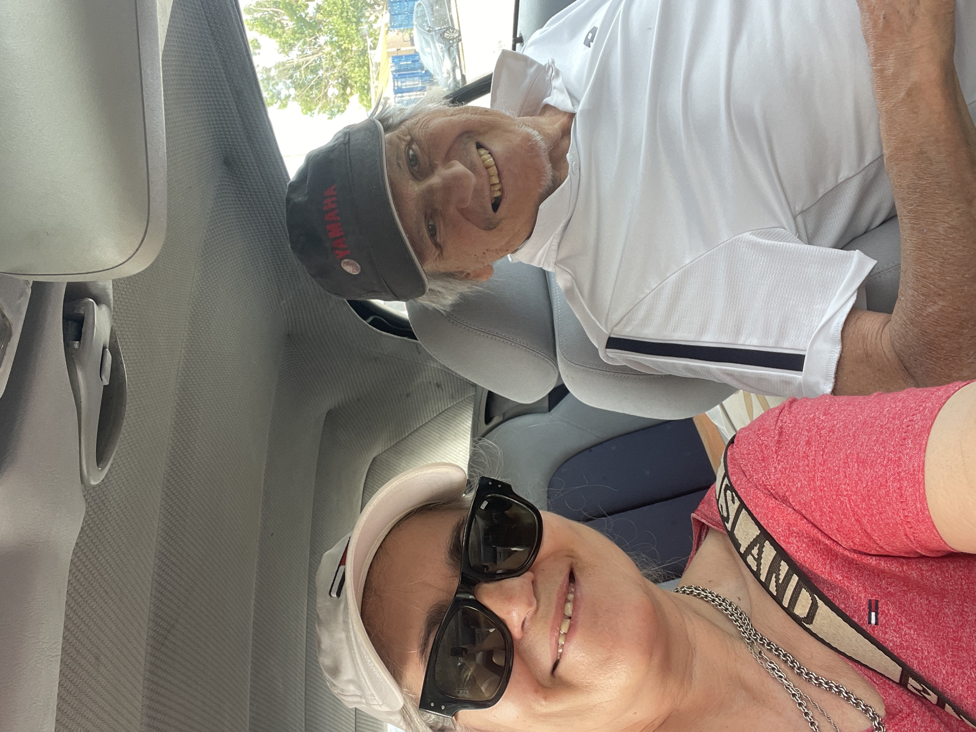 Two people are seated in the passenger section of a car. The person on the left, a woman with graying hair, wears a white baseball cap, black sunglasses, and a red t-shirt. A beige strap with black lettering crosses her chest. The person on the right, a man with gray hair, wears a dark baseball cap with "YAMAHA" printed in red and a white collared shirt. Both individuals are smiling and looking towards the camera. The car's interior is visible, with textured gray headliner and seats. Outside the car window, trees, a fence, and buildings are visible, suggesting an urban or suburban environment. The image appears to be a selfie, taken from inside the car. The location is identified as Follonica, Italy.