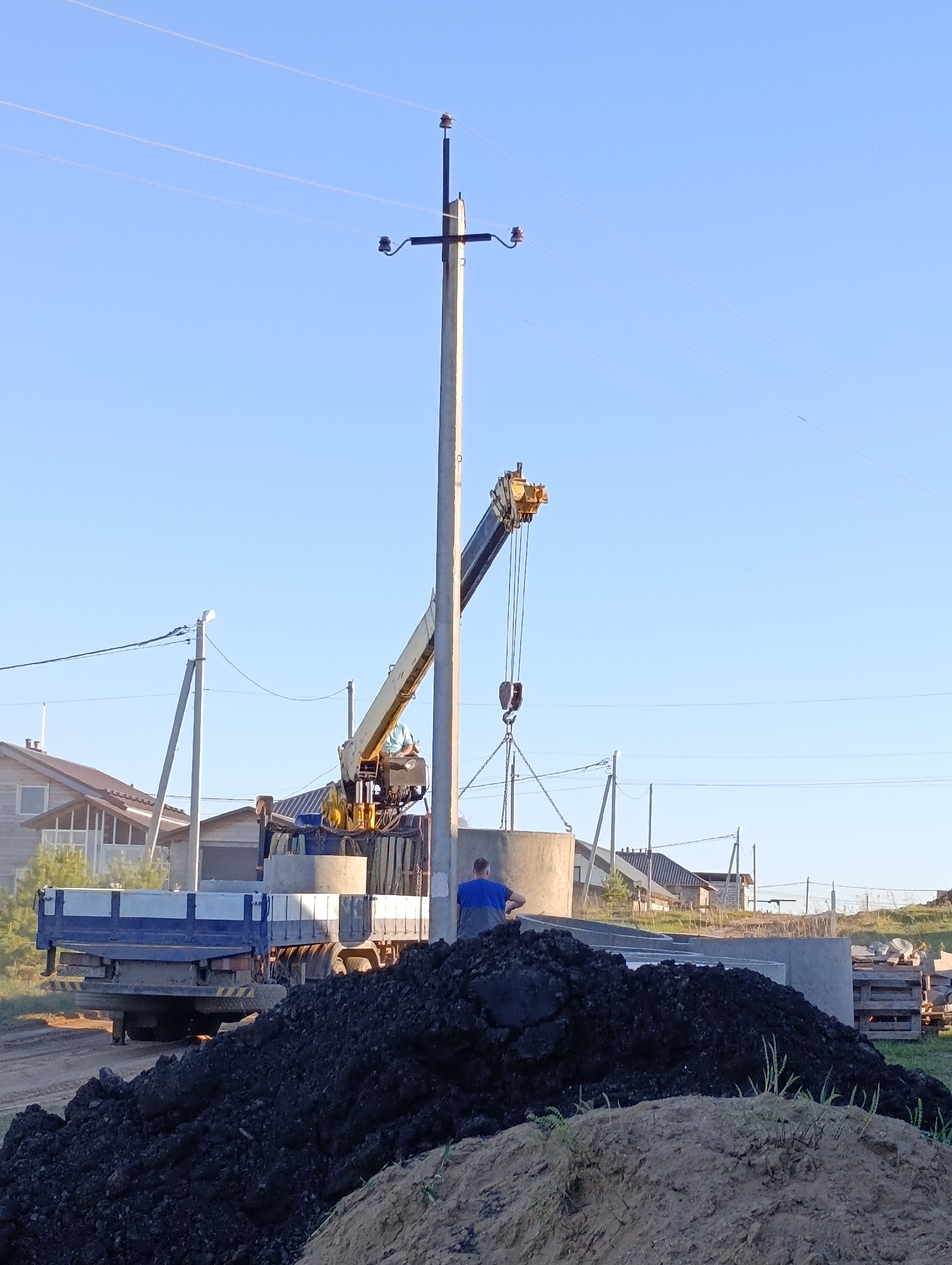 The image depicts a construction site in Chaikovsky, Russia. A truck with a crane is lifting a concrete ring, likely for a well or sewer. A man in a blue shirt stands near the concrete ring. A pile of black dirt is in the foreground, with a smaller pile of tan dirt beneath it. In the background, houses and utility poles are visible under a clear, blue sky.