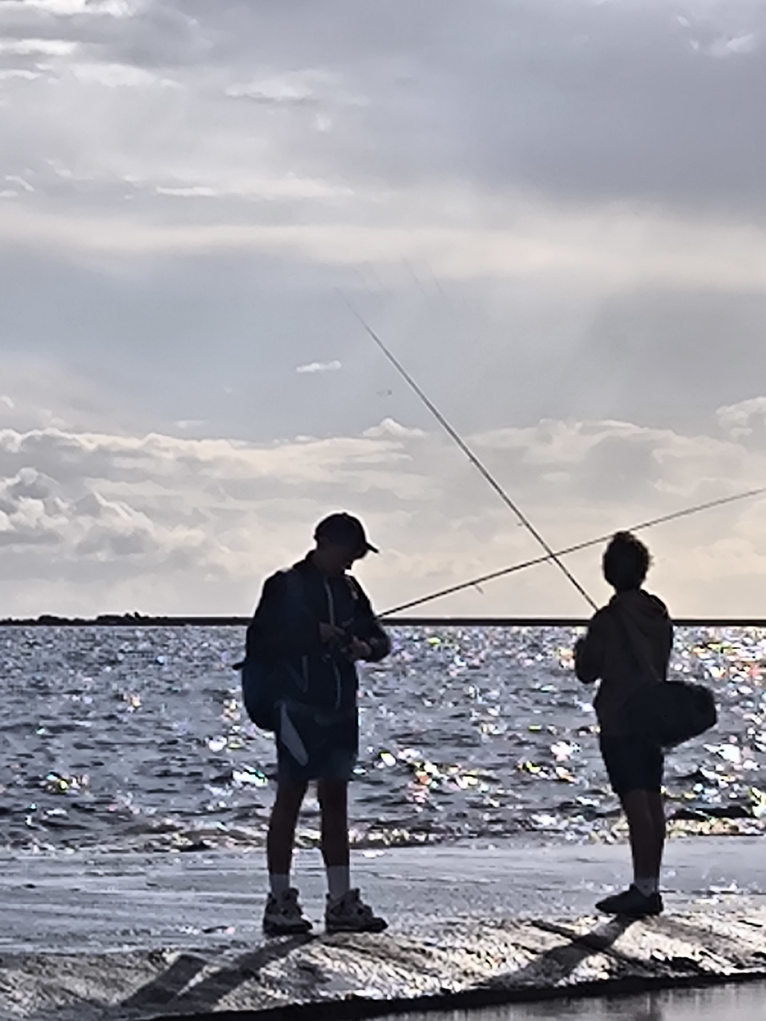 Two silhouetted figures, likely young males, are standing on a concrete or stone structure near the water's edge, engaged in fishing. The figure on the left, wearing a cap, jacket, and shorts, is holding a fishing rod and appears to be attending to something in their hands. The figure on the right, wearing a hoodie and shorts, also holds a fishing rod and a bag is slung over their shoulder. The water behind them is choppy and reflects the light, suggesting a sunny or partly cloudy day. The sky is filled with dramatic clouds. The overall scene is backlit, creating strong silhouettes. Given the context of fishing and likely urban waterfront, this could be a scene from the Daugava River in Riga, Latvia, possibly near a pier or embankment.