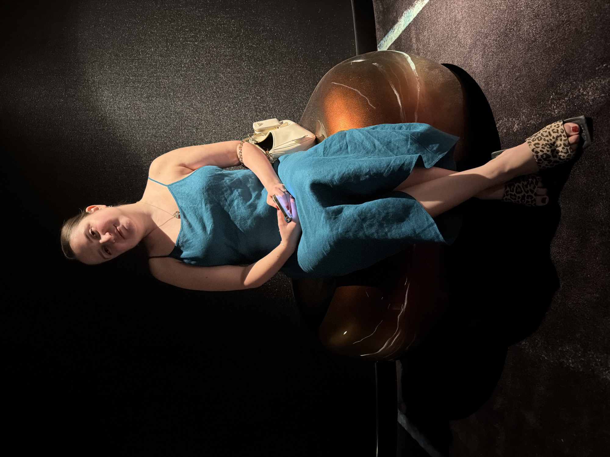 A medium-shot, slightly high-angle, full-body portrait shows a young woman with medium-toned skin and dark hair pulled back, sitting on a rounded, metallic-bronze bench. She is looking towards the camera and smiling slightly. She is wearing a teal, spaghetti-strap midi dress, a silver pendant necklace, and leopard-print slides with square toes. Her left hand rests on her lap holding a smartphone, while her right arm is bent, resting on her leg. A small, cream-colored crossbody bag with gold hardware is slung over her right shoulder. The background is dark, with a textured black wall to the left and a hint of a dark, carpeted floor with subtle lighting patterns to the right. The overall lighting is dim and dramatic, with a spotlight illuminating the woman and the bench. The setting is indoors, possibly a dimly lit lounge or modern venue in Dubai, United Arab Emirates.