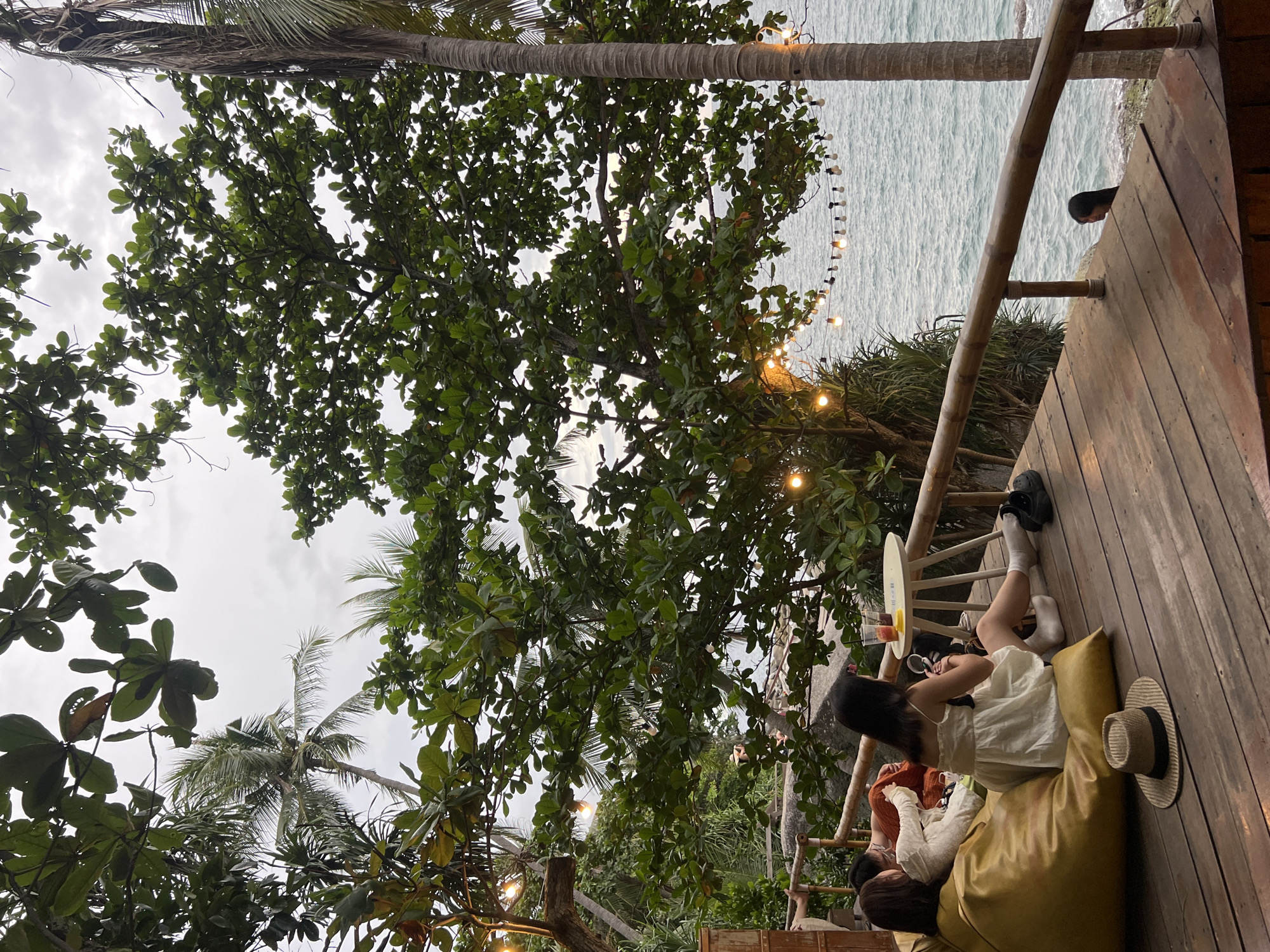 The image depicts a group of people relaxing on a wooden deck overlooking the ocean in Karon, Thailand. In the foreground, two individuals are seated on a large yellow beanbag chair. One person, wearing a white dress and white socks, is propped up on their elbow, appearing to be talking or interacting with someone beside them. A straw hat with a dark band rests on the beanbag next to them. Another person, wearing a light-colored long-sleeved shirt, is partially visible next to them, also appearing relaxed.
Further back on the deck, another person is leaning against a wooden railing, looking out at the ocean. A string of festive lights hangs from a bamboo pole above them, suggesting it might be late afternoon or early evening. The ocean is calm with gentle waves, and the sky is overcast, indicating potentially cloudy weather. Lush green trees and palm fronds frame the scene, adding to the tropical ambiance. The deck itself is made of weathered wooden planks, with bamboo railings and supports. A small white table with drinks is visible near the person leaning on the railing. The overall atmosphere is one of leisure and relaxation in a beautiful coastal setting.