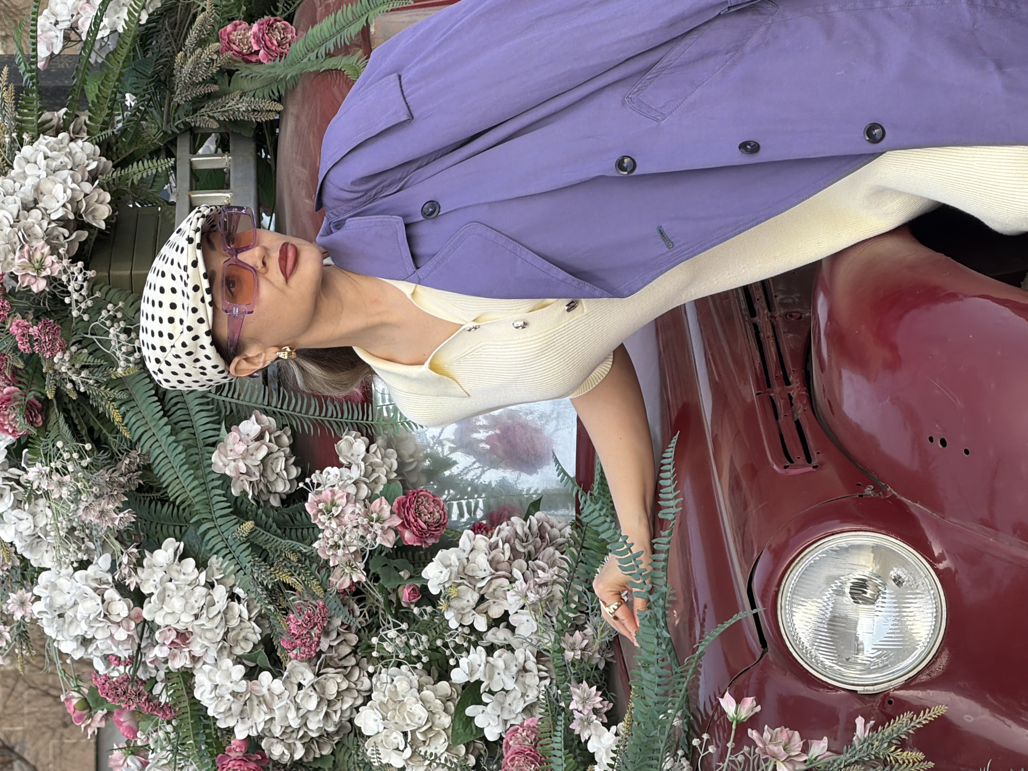 A woman with a polka dot headscarf and purple sunglasses lies on the hood of a maroon vehicle surrounded by an abundance of flowers and ferns. She wears a cream-colored top and a light purple jacket. The scene features an old car, likely an antique or vintage model. The location is Krasnoyarsk, Russia.