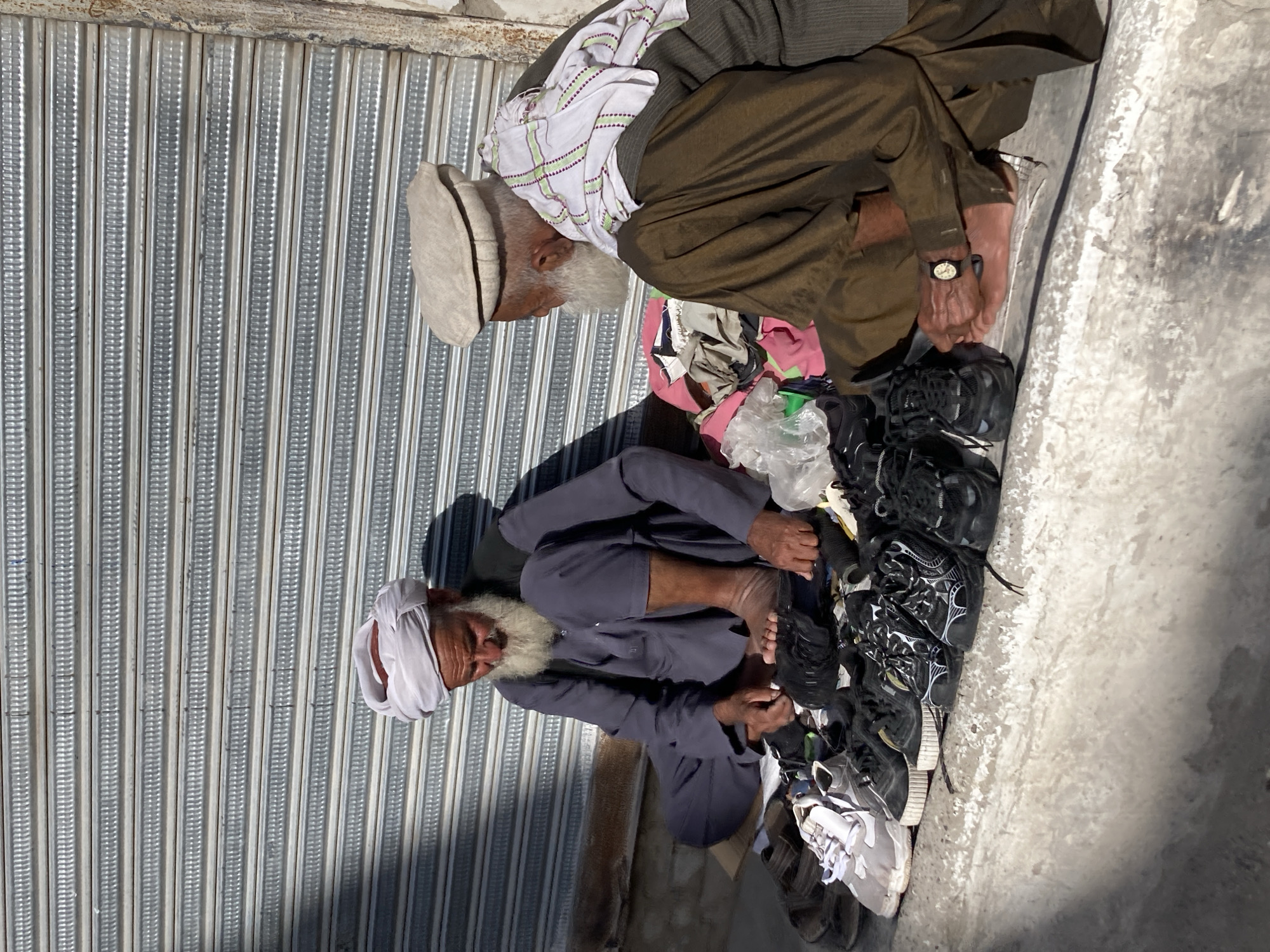 Two older men in traditional Pakistani clothing sit outdoors near a metal corrugated wall, examining shoes. One man wears a grey shalwar kameez and white turban; the other wears a brown shalwar kameez, a white patterned top, and a beige pakol hat. They are surrounded by a pile of shoes of various styles and colors. The environment appears to be a sidewalk or open area within the Quetta City Tehsil of Pakistan.