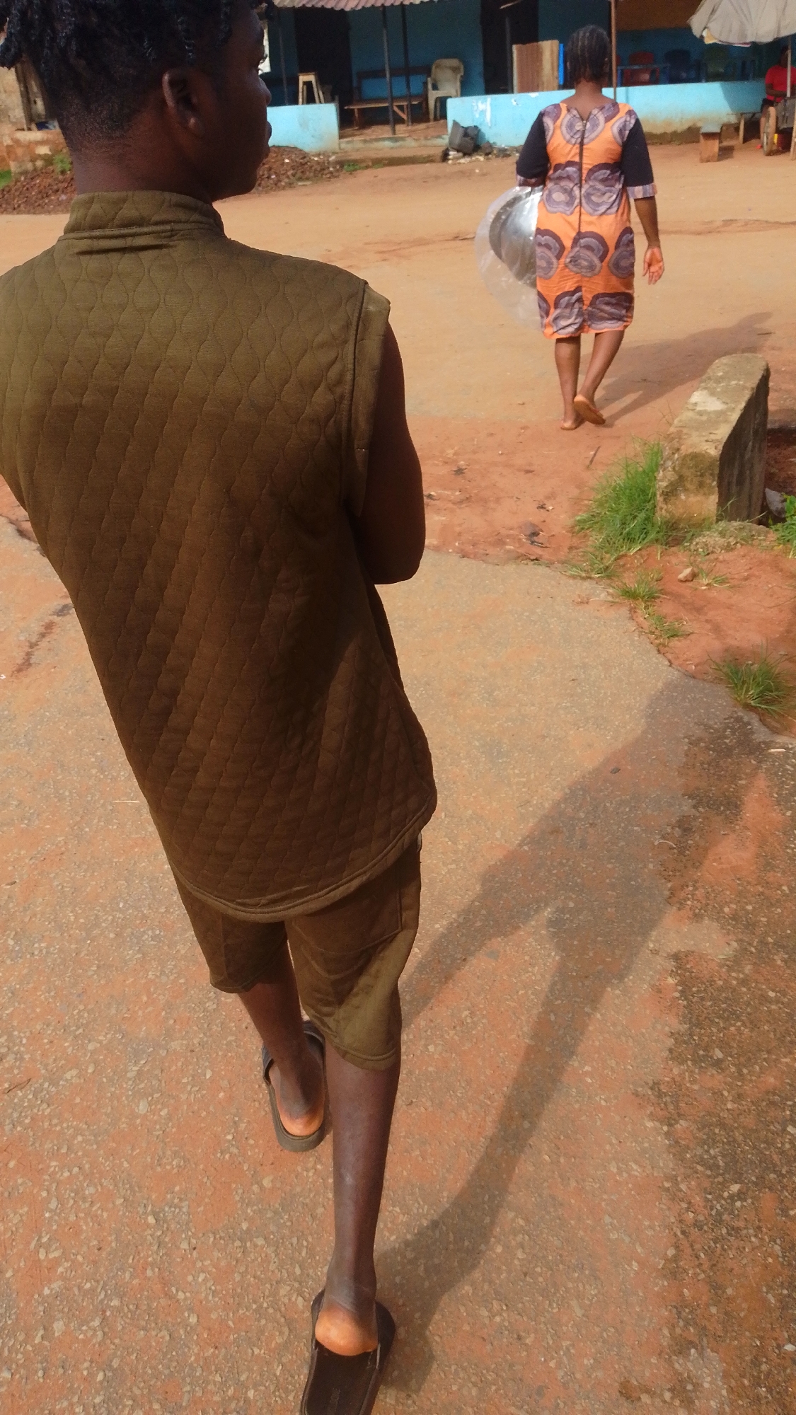 In Enugwu-Ukwu, Nigeria, the photograph shows two individuals walking away from the viewer. The closer person, seen from behind, wears a quilted olive-green sleeveless jacket and matching shorts with brown sandals. The second person is further in the background, carrying a large, circular item in a clear plastic bag. This person is wearing an orange and brown patterned dress. They are walking on a red clay road, partially paved with concrete. In the background is a building with a blue facade.