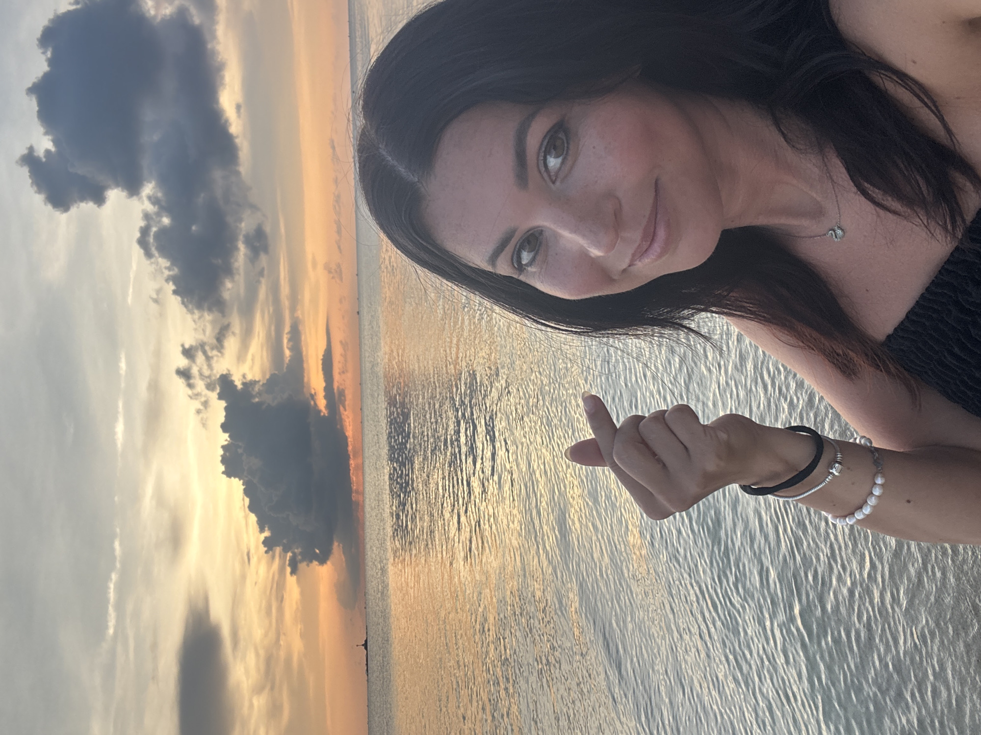 A woman is posing for a selfie at a beach at sunset. The woman is fair-skinned with dark hair and brown eyes, and is wearing a dark-colored top and a silver necklace. Her left arm is outstretched with her fingers pinched together in a "finger heart" gesture. She is wearing two bracelets on her left wrist, one a black elastic band and another of pearls. The background consists of a body of water, the ocean, and a sunset sky. The sun is setting over the water and the sky is orange and yellow near the horizon. There are dark clouds in the sky. Based on the environment and the clothing, it is plausible that the photo was taken at a beach in Baan Sri Thanu, Thailand, as requested.
