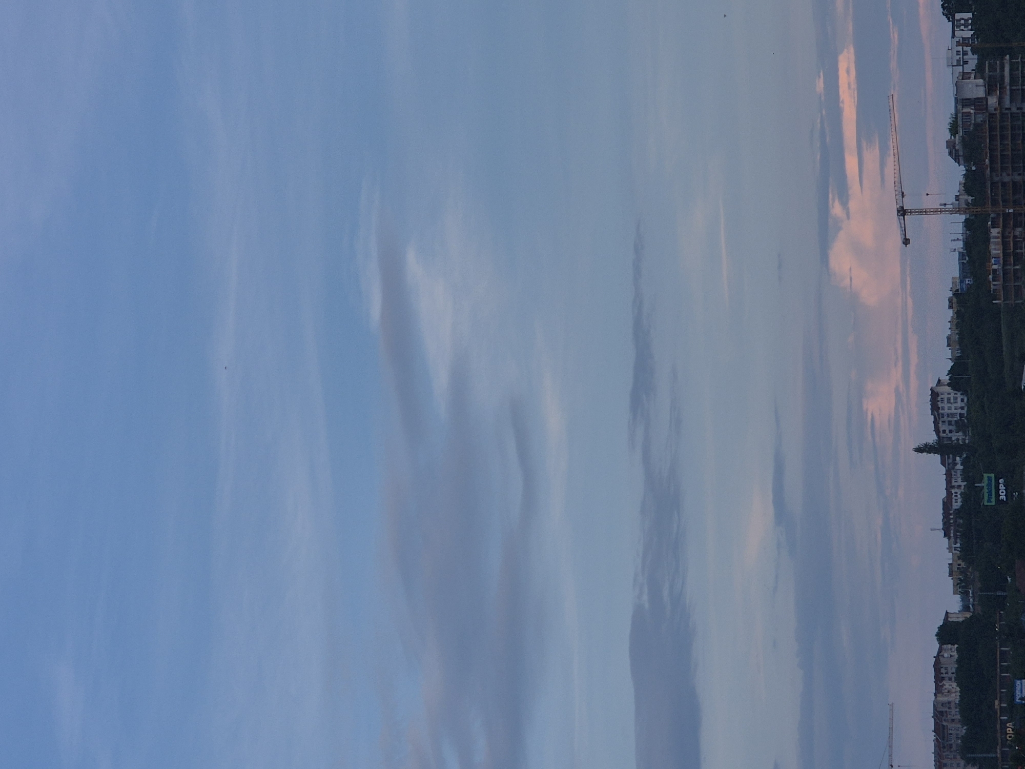 The image displays an urban landscape at either dawn or dusk, characterized by a prominent and colorful sky. The photograph, when correctly oriented, shows a wide expanse of sky occupying the majority of the frame, with the horizon line and city structures visible towards the bottom.
The sky transitions from a light blue at the upper left to warmer hues of pink and orange closer to the horizon, particularly in the middle-right section, indicating a low sun angle. Scattered clouds, some appearing wispy and others in elongated bands, add texture to the sky. A few tiny dark specks, possibly birds, are visible against the blue sky.
Along the horizon, an urban environment unfolds with a mix of completed buildings and structures under construction, evidenced by visible skeletal frames. At least two large construction cranes stand out against the sky, suggesting ongoing development. Dark outlines of trees and vegetation are interspersed among the buildings.
Visible text on green billboards includes "Praktiker" and "ЗОРA" (ZORA), and further to the left, "DECATHLON." A partial sign reads "OPA." The overall scene conveys a sense of calm often associated with sunrise or sunset, set against a backdrop of urban expansion. No specific activity or people are discernible.
