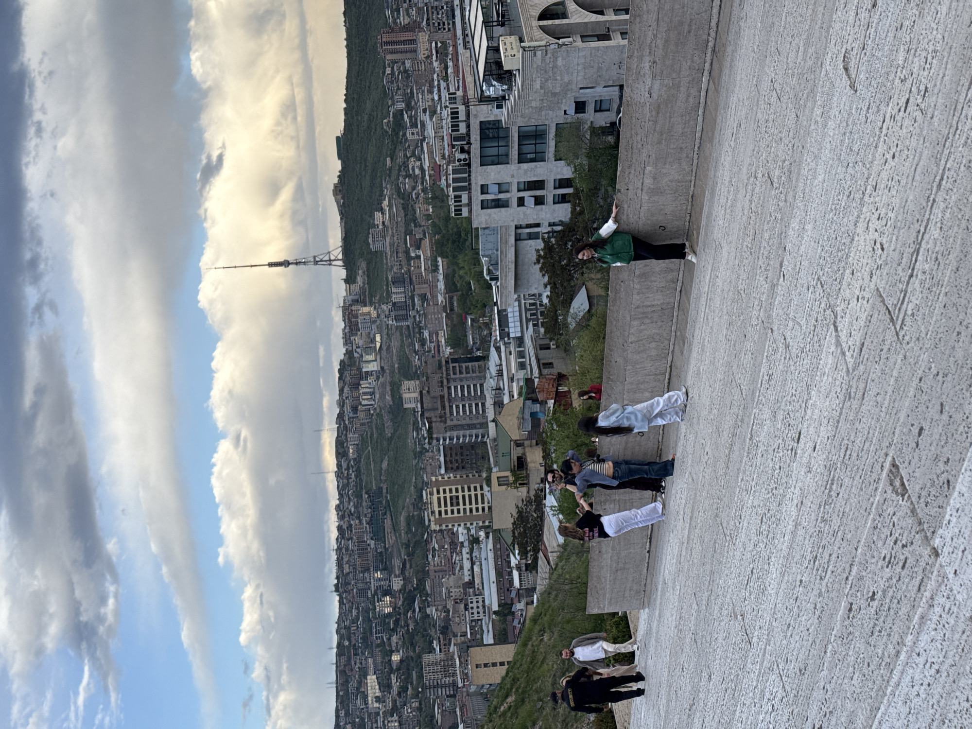 The image captures a scene at the Cascade complex in Yerevan, Armenia. Several people are standing along a concrete barrier or wall, seemingly observing the cityscape. A woman in a green jacket leans against the wall at the top of the image. The background features a dense urban environment with numerous buildings climbing up a hillside, leading to a tree-covered crest. A tall communication tower pierces the sky among the buildings. The sky is partly cloudy with patches of blue visible.