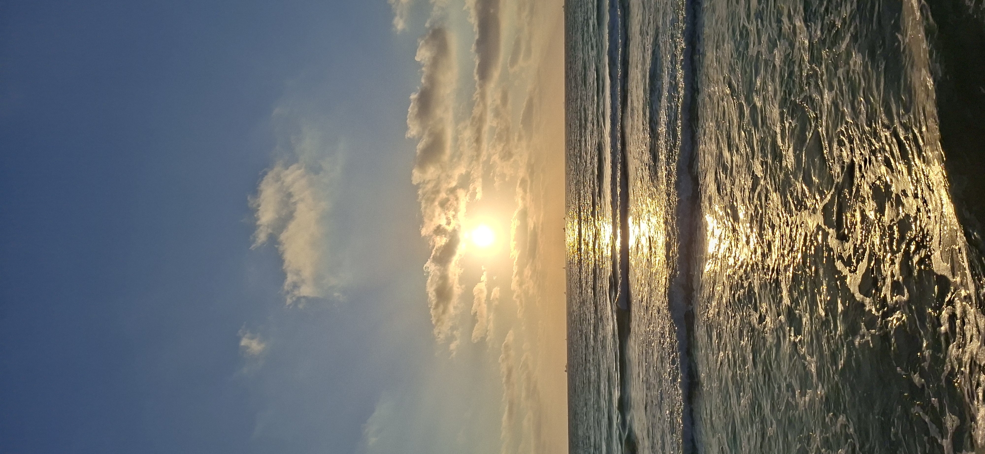 A vertical shot captures a serene ocean scene at sunset. The sky is a gradient of deep blue near the top, transitioning to lighter shades of blue and yellow as it approaches the horizon. Wispy clouds are scattered across the sky, some tinged with the warm, golden light of the setting sun, which is prominently visible as a bright orb partially obscured by clouds.
To the right of the image, the ocean stretches out, with waves gently rolling towards the shore. The surface of the water is illuminated by the sun's reflection, creating a shimmering path of gold across the sea. The waves are not large, suggesting calm conditions, and the water appears to be a deep, rich blue-green. Small white caps are visible on the crests of some waves. The texture of the water's surface is dynamic, with ripples and foam adding depth and visual interest.
The overall mood of the image is peaceful and tranquil, evoking a sense of calm and natural beauty. The time of day is clearly sunset, indicated by the low angle of the sun and the warm, golden light illuminating the clouds and water. There are no visible people or objects in the frame, focusing solely on the natural elements of the sky and sea. No text is present in the image. The location is not specified.