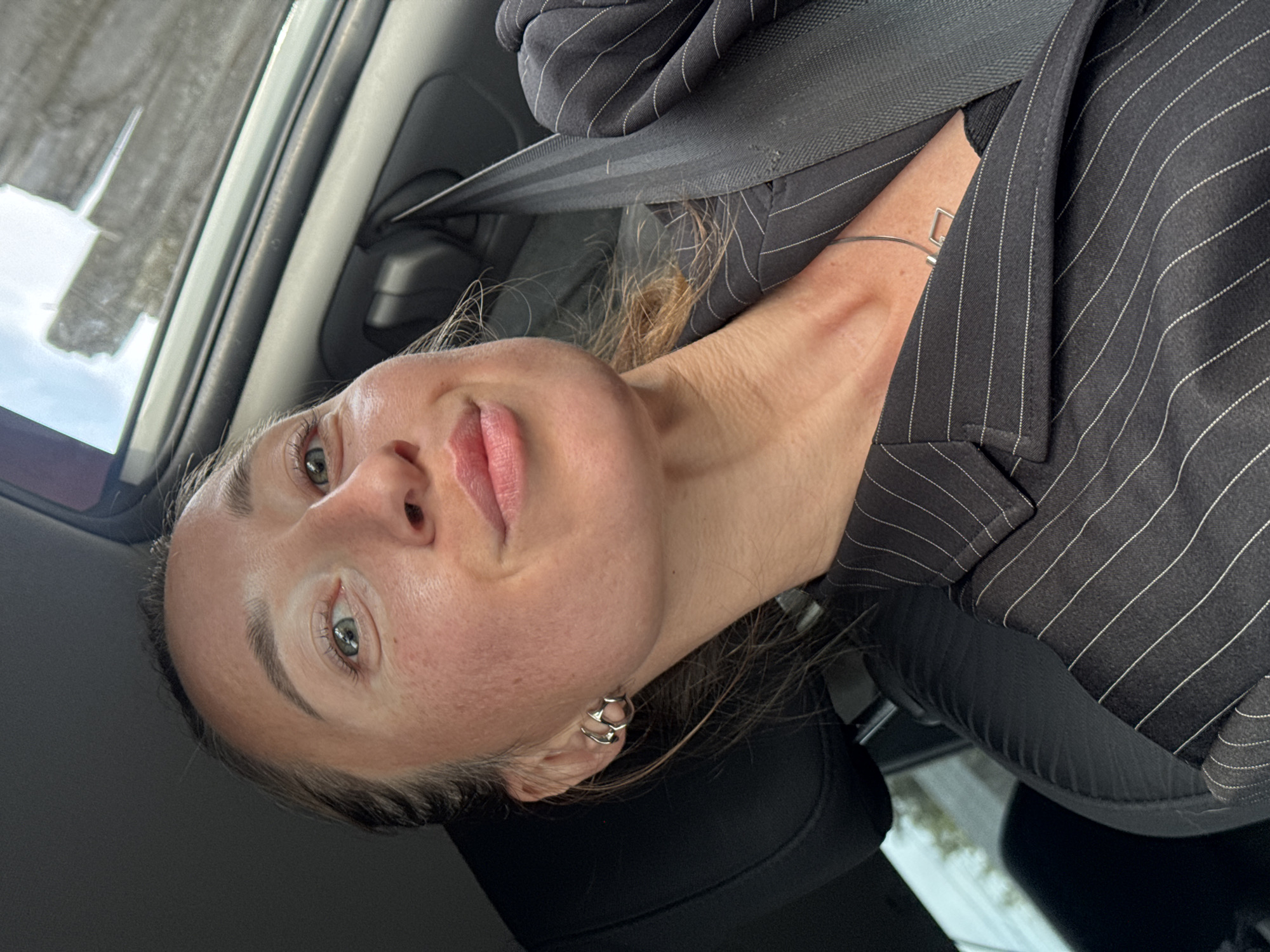 The image is a selfie of a woman wearing a black pinstripe blazer and a seatbelt, seemingly taken inside a car. She has light skin, green eyes, and her hair is pulled back. She is wearing multiple earrings in one ear and a silver bracelet on her wrist. The location is unknown, and there is no verifiable information suggesting the location is Chaikovsky, Russia. There is no additional information to be inferred beyond what is explicitly visible in the image.