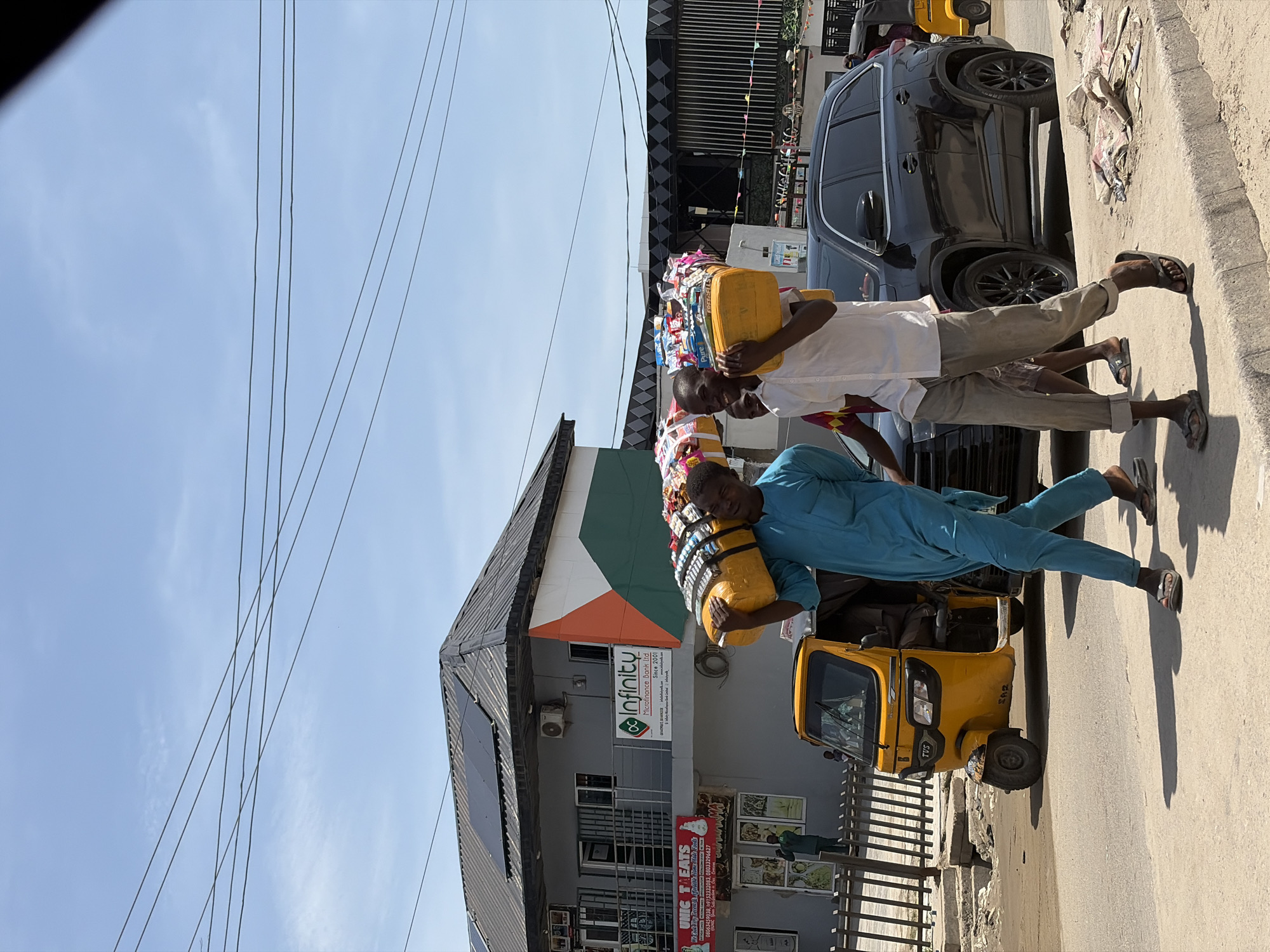 The image depicts a street scene in Ado, Nigeria, featuring two men carrying goods on their heads. One man is wearing a blue outfit and the other a white top with khaki trousers. They are walking along a paved street alongside a yellow "keke" (tricycle taxi) and a black car. The background includes a building with the signage "Infinity Microfinance Bank Ltd" and a food vendor named "Nic Treats". Overhead power lines crisscross the sky. The general environment appears to be a commercial or urban area.