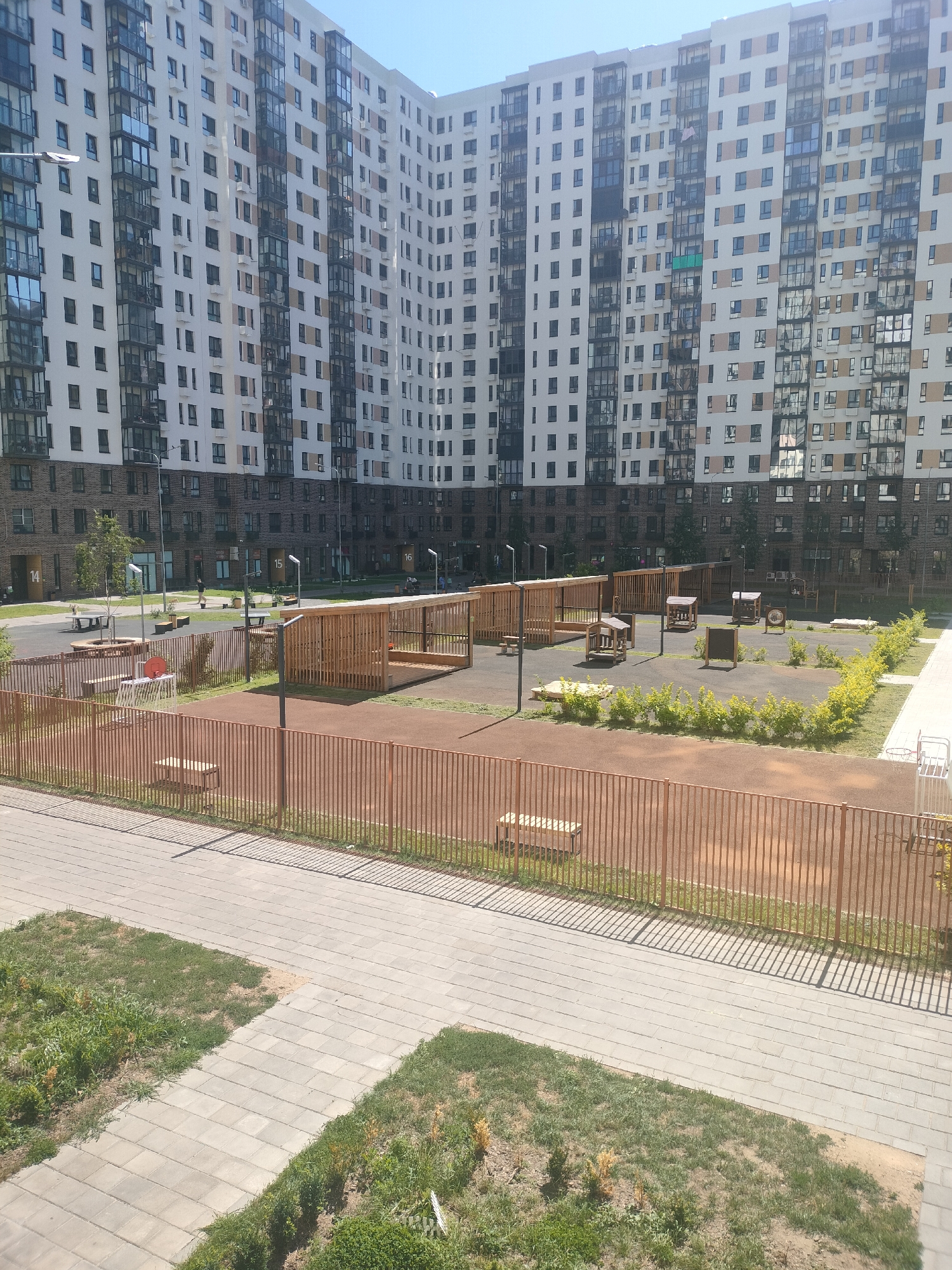 This is an eye-level, wide-angle, outdoor, daytime photograph of a residential courtyard in Lyubertsy, Russia. The scene is dominated by a large, multi-story apartment building with a curved facade, featuring numerous balconies and windows. In the foreground, a paved walkway with grassy patches borders a fenced-off area that appears to be a playground or recreational space. This area contains wooden shelters, benches, a basketball hoop, and some smaller play structures. The fencing is a uniform reddish-brown color. The sky is clear and blue. The overall impression is of a modern, urban residential environment.
