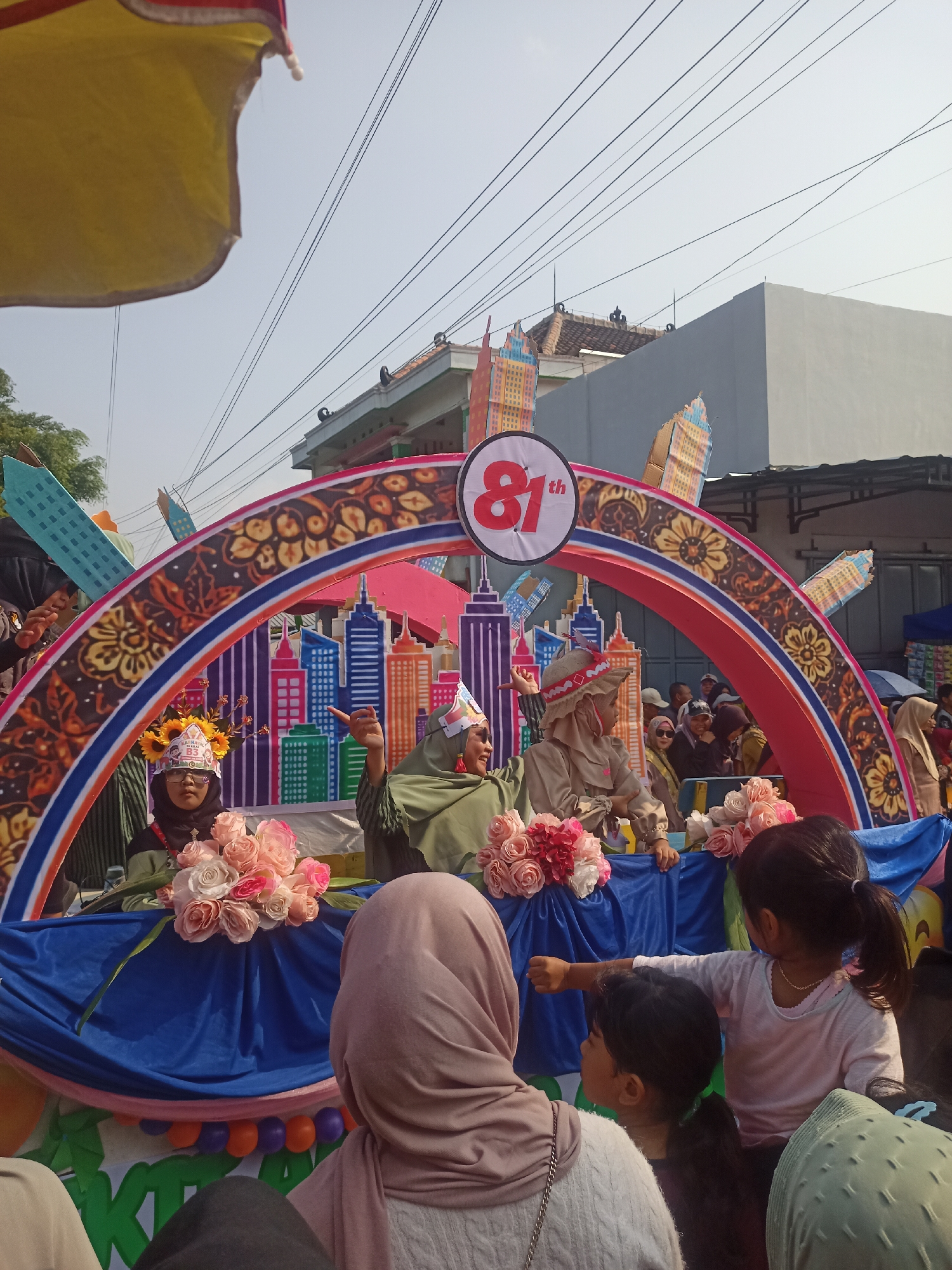 An outdoor daytime scene in Gemolong, Indonesia, captures a festive parade float. The float is adorned with a large archway featuring a stylized cityscape and patterned decorations. On the float, three women in traditional head coverings are visible, with one in the center gesturing. In front of them, a young girl wearing a crown and a sunflower headpiece holds a bouquet of pink and white roses. In the foreground, two young girls, one in a pink shirt and the other in a grey shirt, observe the parade, with the girl in pink pointing. The background reveals buildings and overhead wires, characteristic of an urban environment.