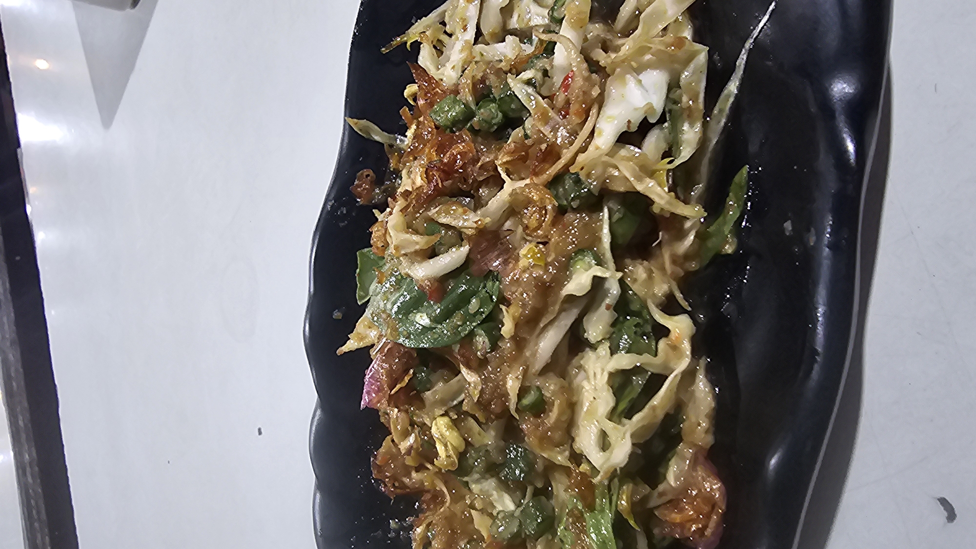 A close-up, overhead shot shows a black, wavy-edged plate filled with a colorful salad. The salad appears to be a mix of shredded cabbage, green beans, red onion, and herbs, all tossed in a dark, savory dressing. Crispy fried shallots are sprinkled generously on top, adding a textural contrast. The plate sits on a white, speckled surface, and a dark band with reflective lights is visible at the top of the frame, suggesting an indoor setting, possibly a restaurant or food stall. The overall impression is one of a vibrant and appetizing dish. The location is Pasirhalang, Indonesia.