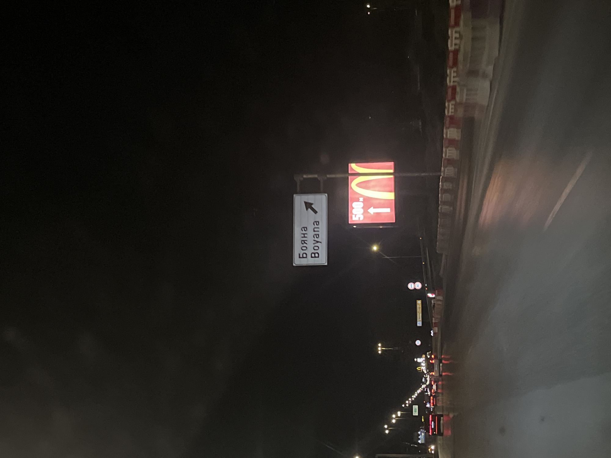 The image shows a nighttime street scene on a road in Sofia, Bulgaria. A road sign indicates the direction to Boyana with an arrow pointing to the left. Another sign shows a McDonald's logo and indicates it is 500 meters ahead. Streetlights illuminate the road and oncoming traffic. The scene appears to be captured from a moving vehicle.