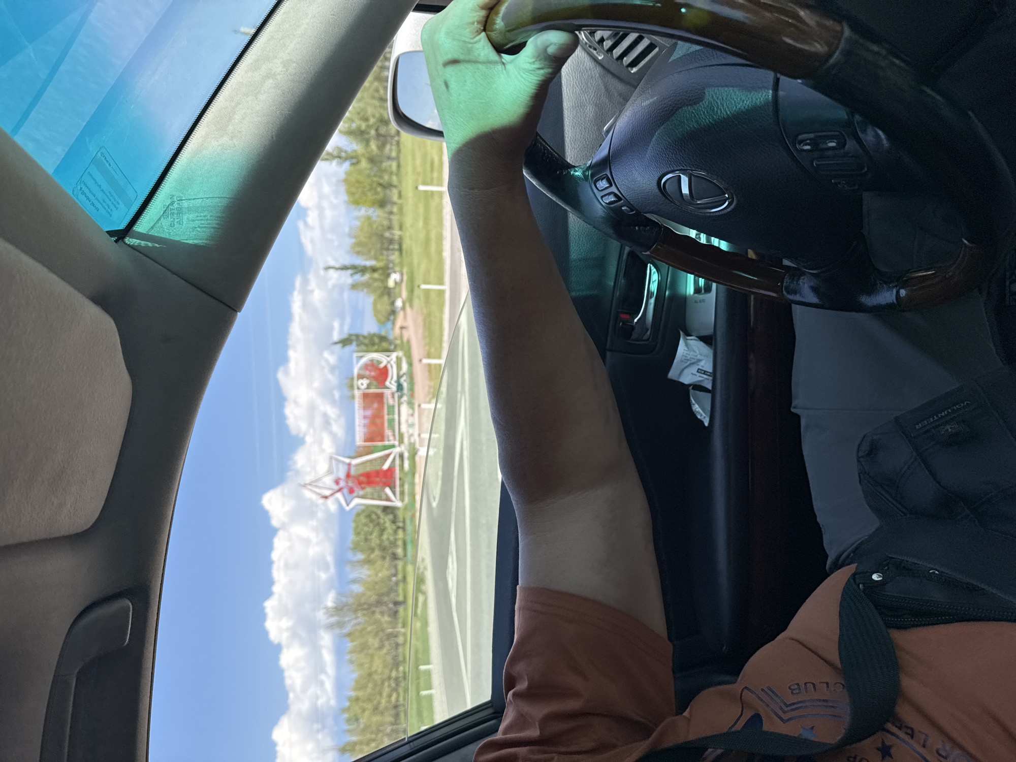 An individual is driving a Lexus sedan in Balykchy, Kyrgyzstan. The exterior view through the windshield shows a large red star monument with scaffolding, surrounded by trees and a clear blue sky with white clouds. The driver's arm and hand are visible resting on the steering wheel, with a green light filtering through the windshield onto their skin. The interior of the car features a wooden-accented steering wheel and dashboard. The scene suggests a daytime drive in a scenic, possibly rural or park-like area of the city.