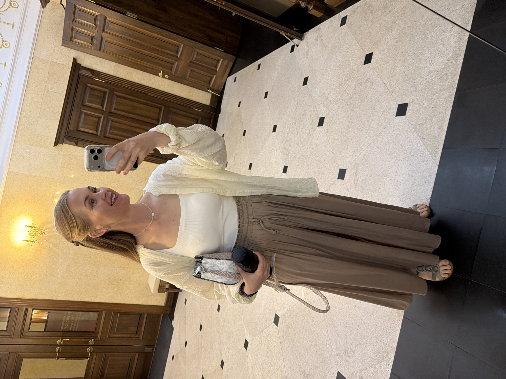 A woman is taking a selfie in what appears to be the lobby of a building with ornate wooden doors and a marble floor. She is wearing a white tank top, a cream-colored cardigan, and a long, flowing brown skirt. She is holding a purse and a water bottle. The woman has blonde hair and is smiling. The location is stated to be in Khabarovsk, Russia.