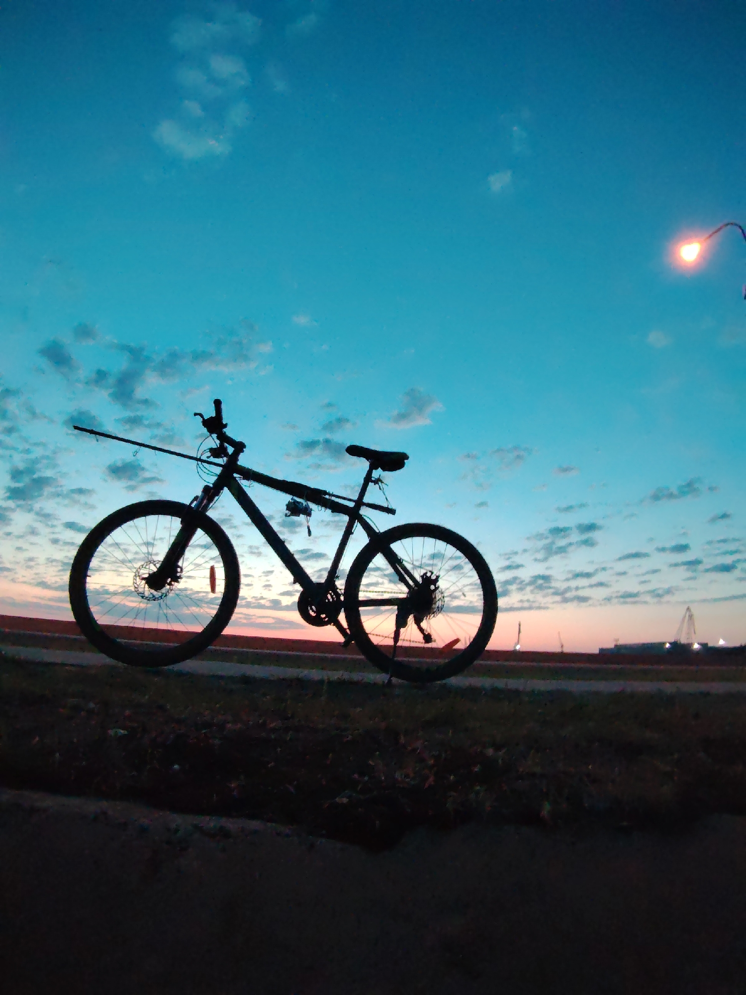 A bicycle with a fishing rod attached is silhouetted against a blue and orange sky at sunset or sunrise. The bike is on a patch of grass next to a paved surface. In the distance, there are structures that appear to be industrial or harbor-related, possibly cranes or large buildings. A streetlight is visible in the upper right corner. Based on the context, this image could have been taken in Chaikovsky, Russia. The objects include: a bicycle, a fishing rod, the ground/pavement, streetlight and industrial structure. No people are in view in the image. The actions/interactions include: a fishing rod attached to the bicycle for travel.