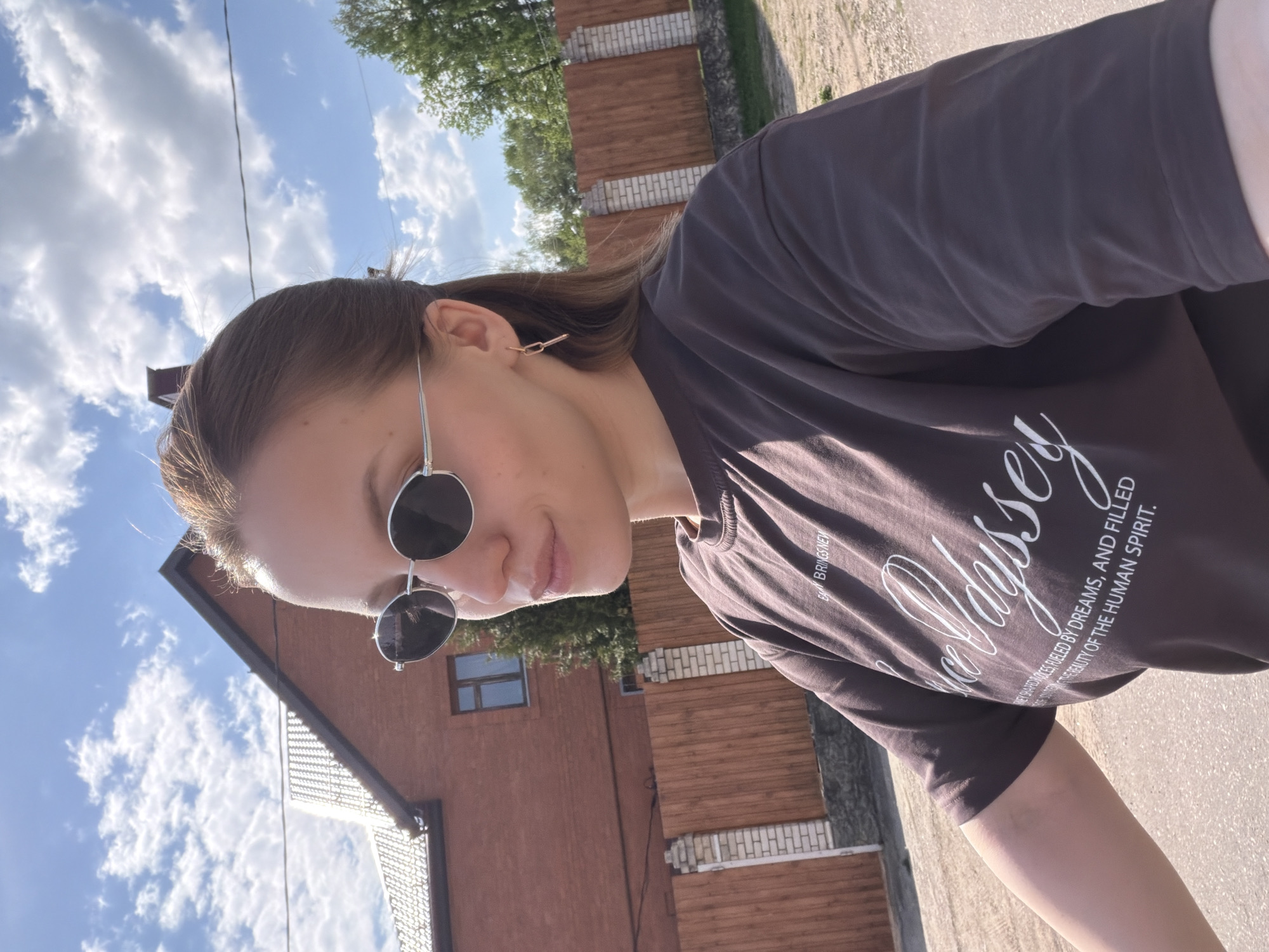 A young woman wearing round sunglasses and a brown t-shirt is outdoors, presumably in the city of Reshetnikovo, Russia. The shirt has text that reads "EVERY BRINGS NEW" and "EASE ODYSSEY NEW SHAPED VOICES, FUELED BY DREAMS, AND FILLED THE BEAUTY OF THE HUMAN SPIRIT." Behind the woman, there is a house with a brown roof and walls, and a partially visible wooden fence. The sky is blue with scattered white clouds. The photo appears to be taken from a low angle.