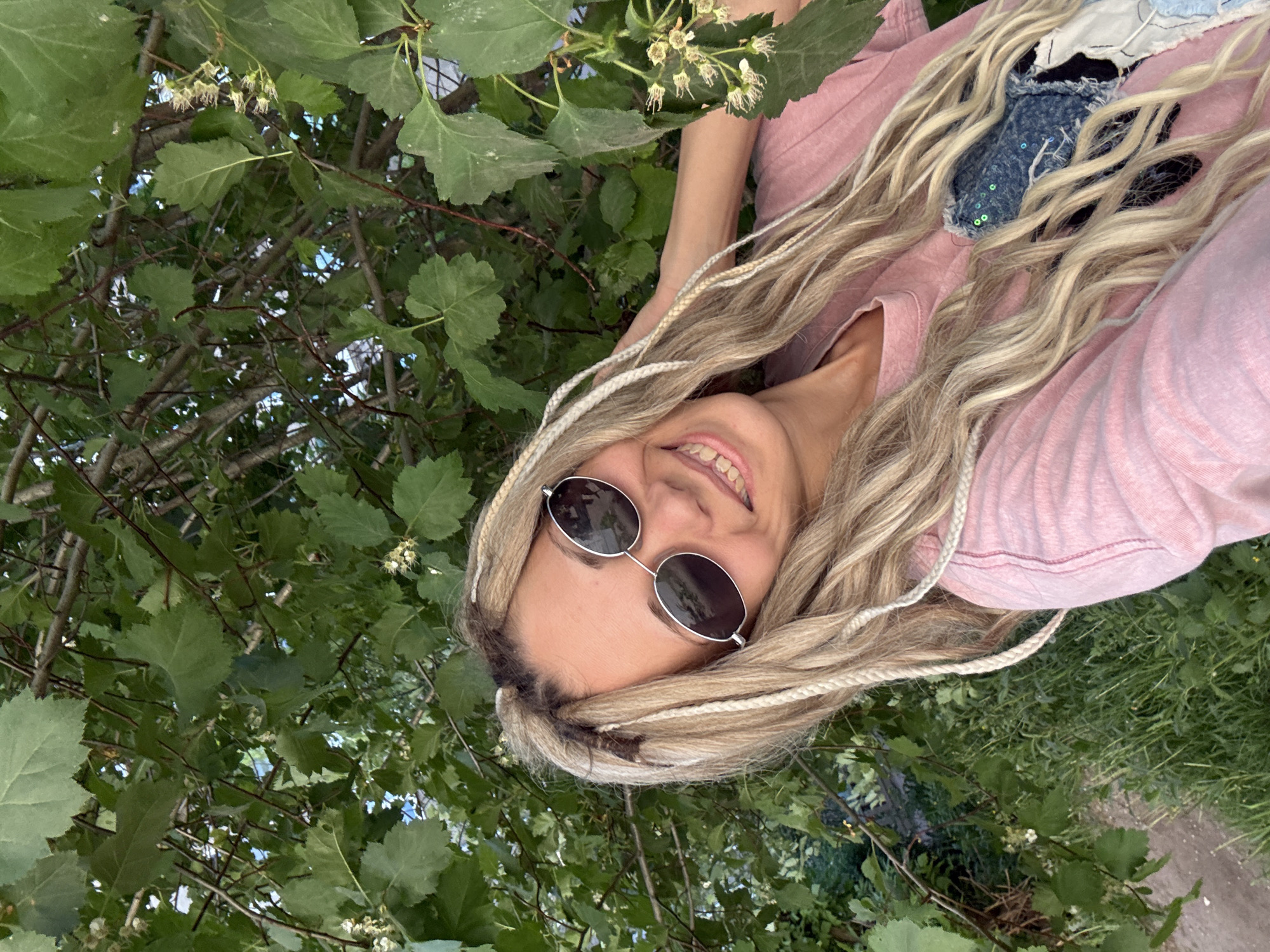 A smiling woman wearing round sunglasses and a pink top is positioned amongst green foliage, possibly a tree or bush. Her blonde hair is braided with white strands. The setting appears to be outdoors, possibly in the Rusanovo area of Russia.