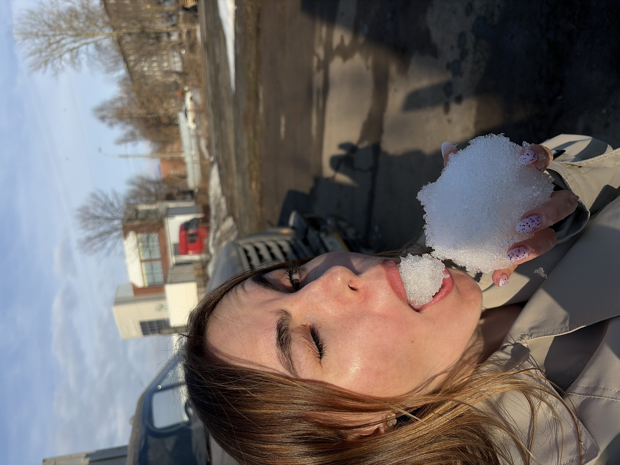 A woman is holding and eating a chunk of snow. The background shows a mix of urban and natural environments, with buildings and bare trees visible. The sky is blue with some clouds. The woman's hand holding the snow features patterned nail polish. The location appears to be outdoors, possibly a street in a city based on the surrounding architecture. Based on the location as Izhevsk, Russia, it can be inferred that this scene is taking place in the spring when there is still snow on the ground.