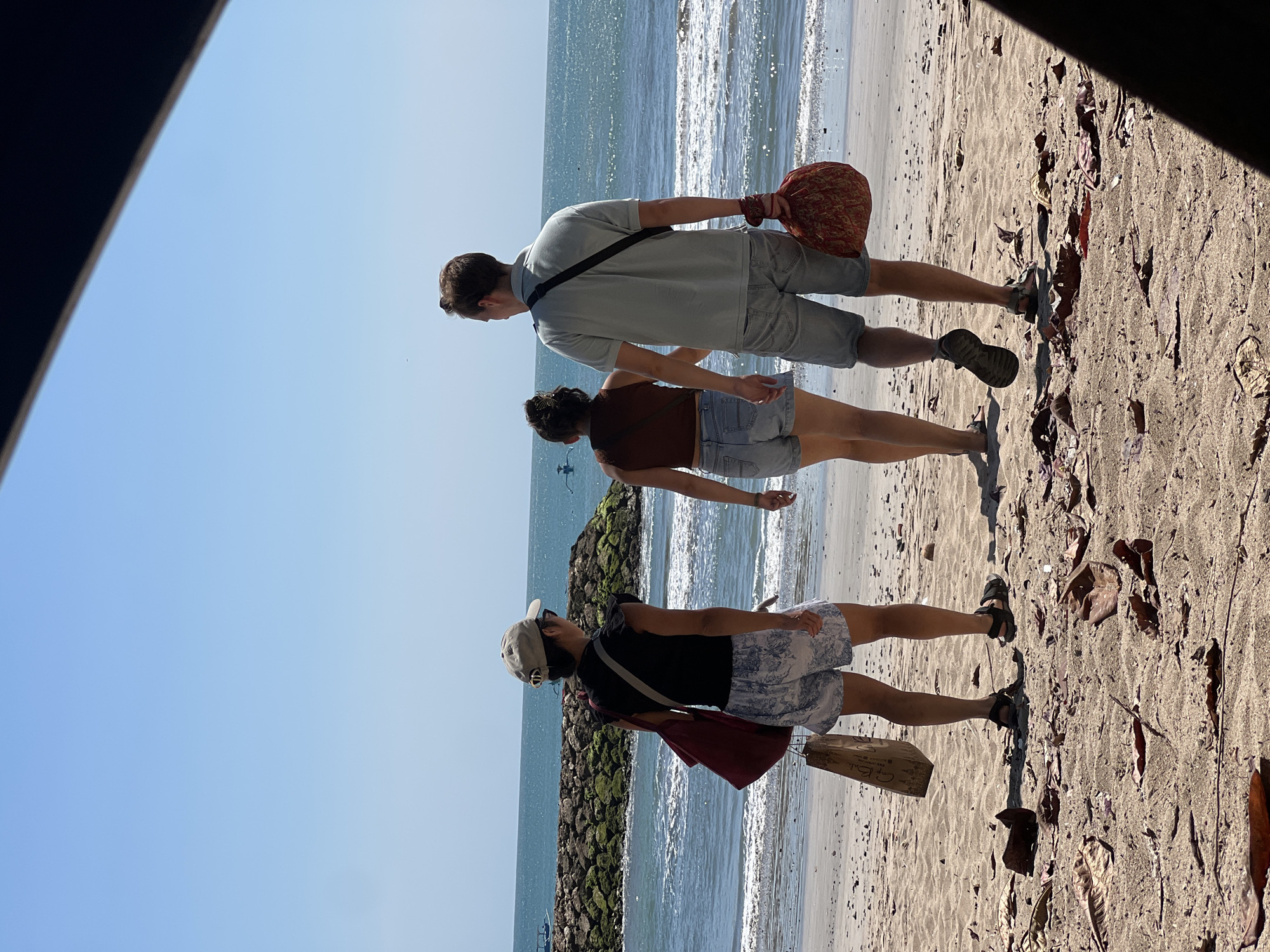 Three individuals are walking on a sandy beach in Kuta, Indonesia, facing away from the camera towards the ocean. The person on the left, wearing a light-colored baseball cap, black t-shirt, and patterned shorts, carries a maroon tote bag on their left shoulder and a brown paper bag in their right hand. The person in the middle, with dark hair, wears a brown crop top and denim shorts. The person on the right, a male wearing a light blue polo shirt and gray shorts, has a black strap across his chest and carries a red patterned bag in his right hand. The beach has scattered dry leaves and debris. In the background, a stone breakwater with green vegetation separates the sand from the blue ocean, where gentle waves are breaking. The sky is clear and blue.