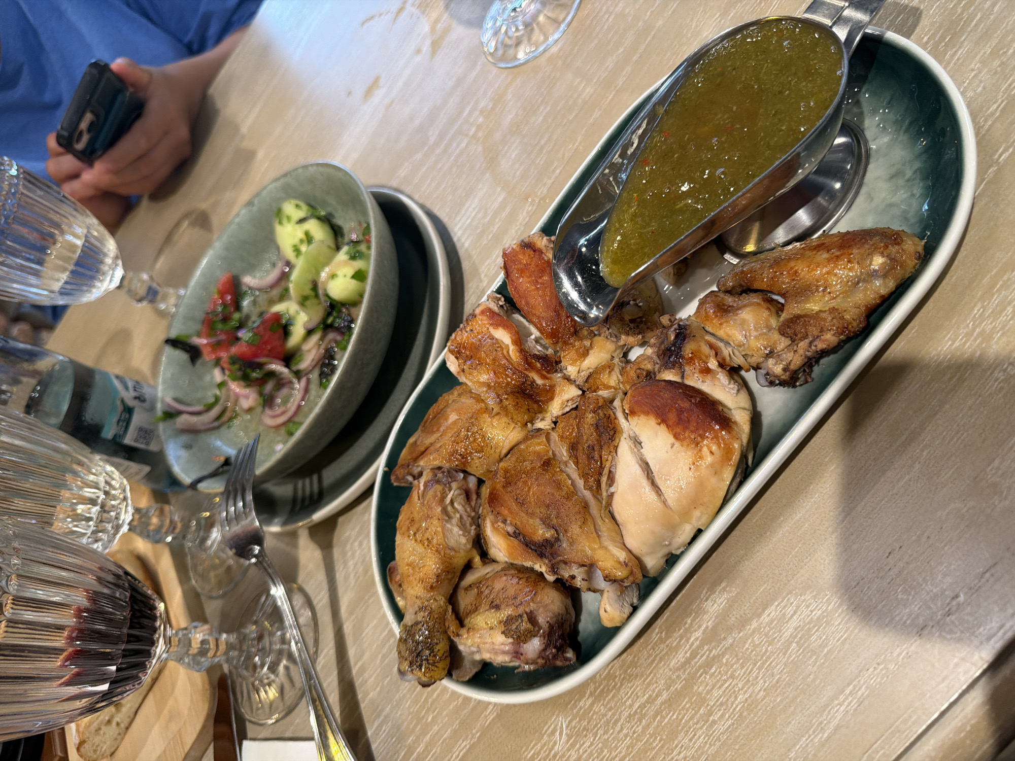 The image shows a dining table at a restaurant. In the foreground, a platter of roasted chicken is served with a green sauce in a metal gravy boat. Behind the chicken, a salad with tomatoes, cucumbers, and red onions is in a green bowl. There are wine glasses and a bottle of water on the table. The table is made of wood. The setting appears to be a restaurant in Tbilisi, Georgia, with the overall ambiance suggesting a casual dining experience.