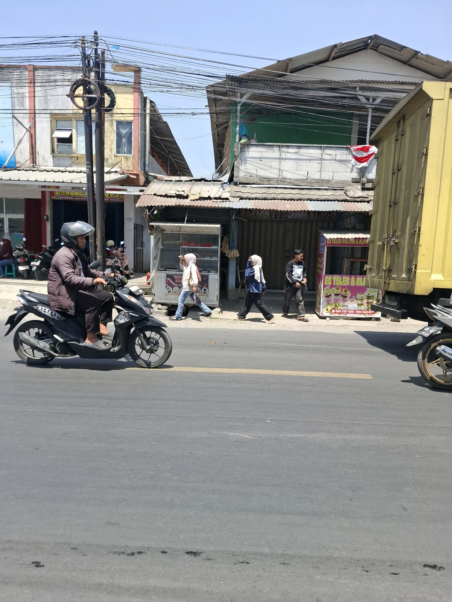 The image depicts a street scene in Sukalarang, Indonesia. In the foreground, a man wearing a helmet and a dark jacket is riding a black motorcycle. He is on the left side of the frame, heading towards the right. The road surface is asphalt with a yellow line marking the lane. In the mid-ground, there are several buildings and a yellow truck on the right. A woman wearing a hijab and a blue top is walking from right to left, carrying a bag. Another man in a black shirt and dark pants is standing near a food stall. The food stall has colorful advertisements for "ES TELOR SULTAN & ALP...". There are also other buildings with corrugated metal roofs and visible electrical wires. Several parked motorcycles are visible in front of one of the buildings. The sky is clear and blue.