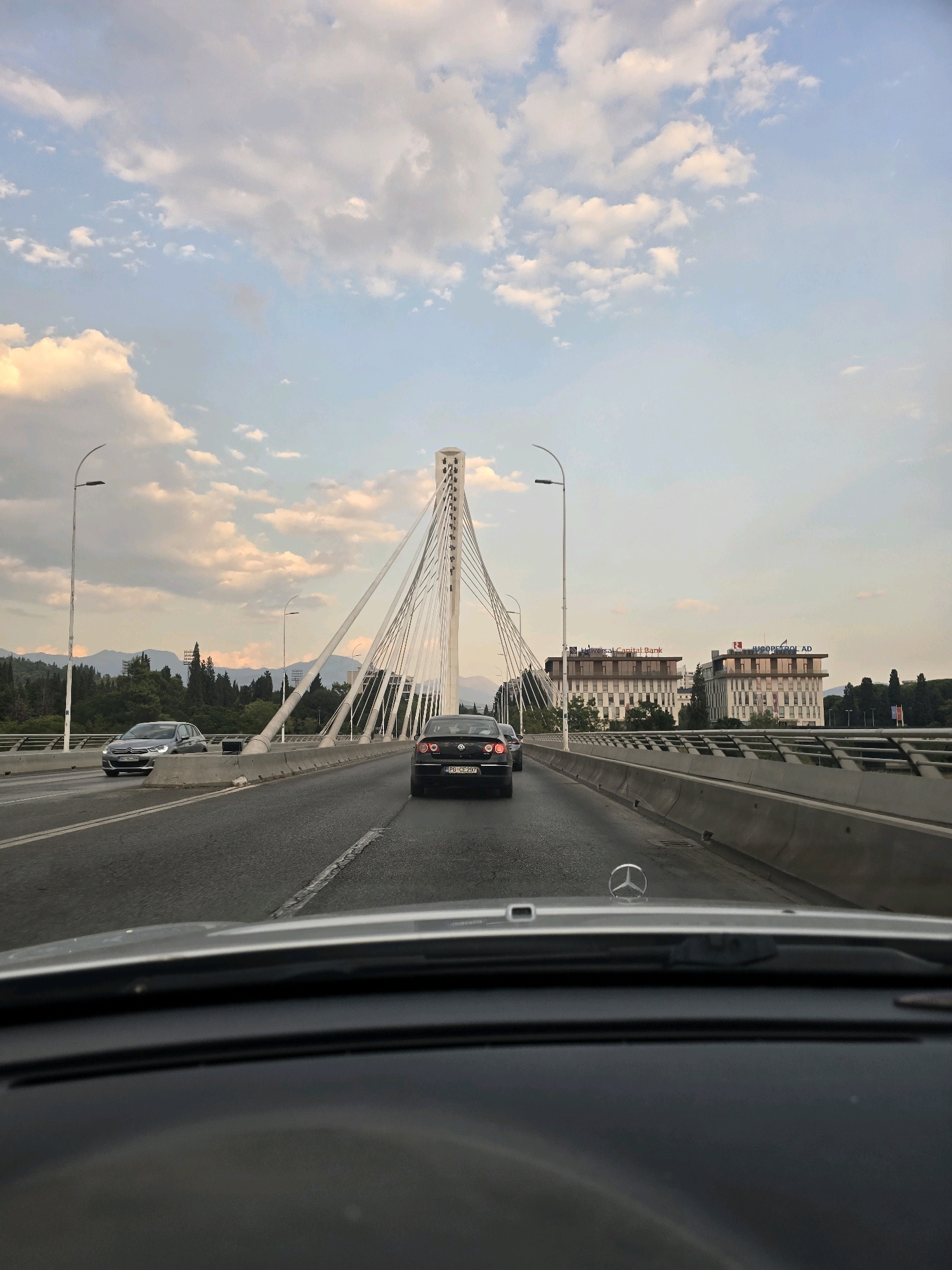 A driver's-eye view from inside a Mercedes-Benz automobile traveling on a multi-lane bridge over a road, with the Millennium Bridge in Podgorica, Montenegro, visible in the background. The sky is filled with scattered clouds, some tinged with the warm colors of sunset or sunrise. The bridge's distinctive cable-stayed design is prominent, with white cables fanning out from a tall central pylon. Two buildings with modern architecture and signage are situated on the right side of the road, one reading "Universal Capital Bank" and the other "JUGOpetrol AD". A gray sedan is in the left lane, and a dark sedan is in the right lane. Mountains and lush green trees form the backdrop beyond the bridge. The foreground shows the car's dashboard and windshield.