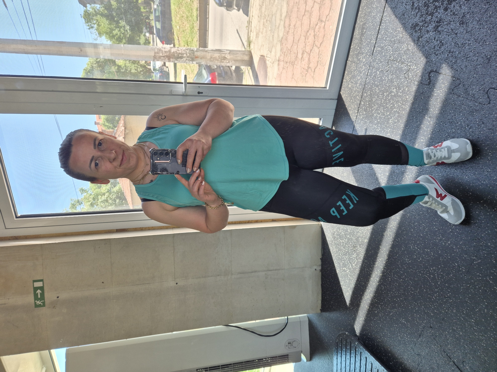 A woman with her hair pulled back and wearing a teal tank top and black leggings with teal accents is taking a mirror selfie in what appears to be a gym. She is holding a smartphone in her right hand. The background shows a large window with a view of trees and a street outside, and part of a gym floor. An exit sign is visible on the wall to the left.