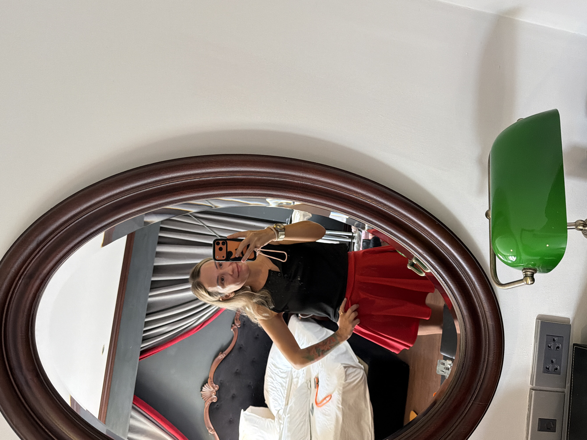 A woman is taking a mirror selfie in a hotel room. She is wearing a black tank top and a red skirt. She has blonde hair and is smiling. In the background, there is a bed with white sheets and a dark headboard. There are also curtains and a mirror. A green desk lamp is visible in the foreground. The room appears to be in a hotel in Thuận Hóa Ward, Vietnam.