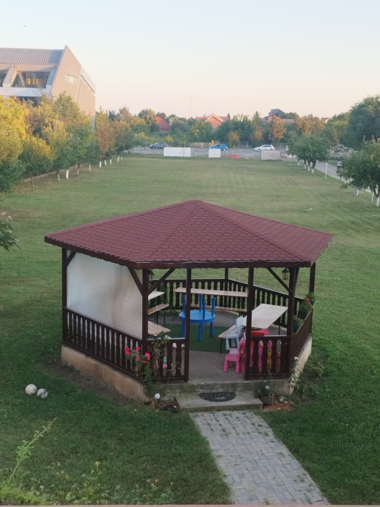This image depicts an outdoor scene with a gazebo as the central focus. The gazebo has a dark red, shingled roof and wooden support posts. Inside, there are several picnic tables, some with benches, and brightly colored children's chairs, suggesting it's a recreational area. A white, translucent screen is visible on one side of the gazebo.
The gazebo is situated on a well-maintained green lawn, with a paved walkway leading to its entrance. Two soccer balls are present on the grass near the gazebo. In the background, there's a row of trees with white-painted trunks, and beyond that, a street with a few cars. Further in the distance, buildings with pitched roofs and a more modern, angled structure are visible, indicative of a mixed urban and suburban setting. The sky is a pale, clear hue, suggesting either early morning or late evening.
This scene is located in Bucharest, Romania, likely in a public park, recreational area, or a residential complex with communal outdoor spaces.