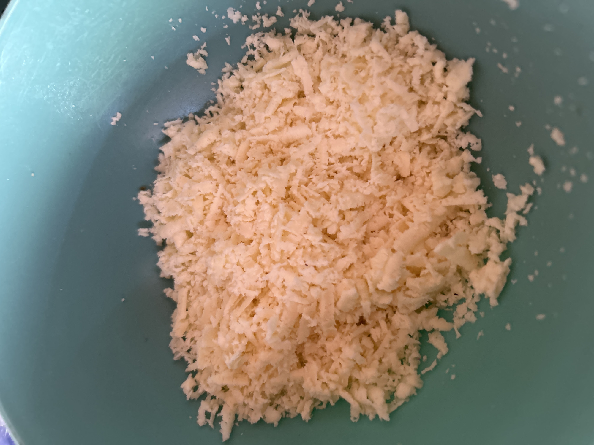 The image shows a close-up, overhead view of finely grated cheese in a light teal bowl. The cheese is a pale yellow color and appears fluffy. The bowl is made of plastic and has a matte finish. The lighting is warm, suggesting it might be artificial or from a window on a sunny day. There are no people or specific landmarks visible, and the location of Santa Ana, Venezuela is not depicted in the image.