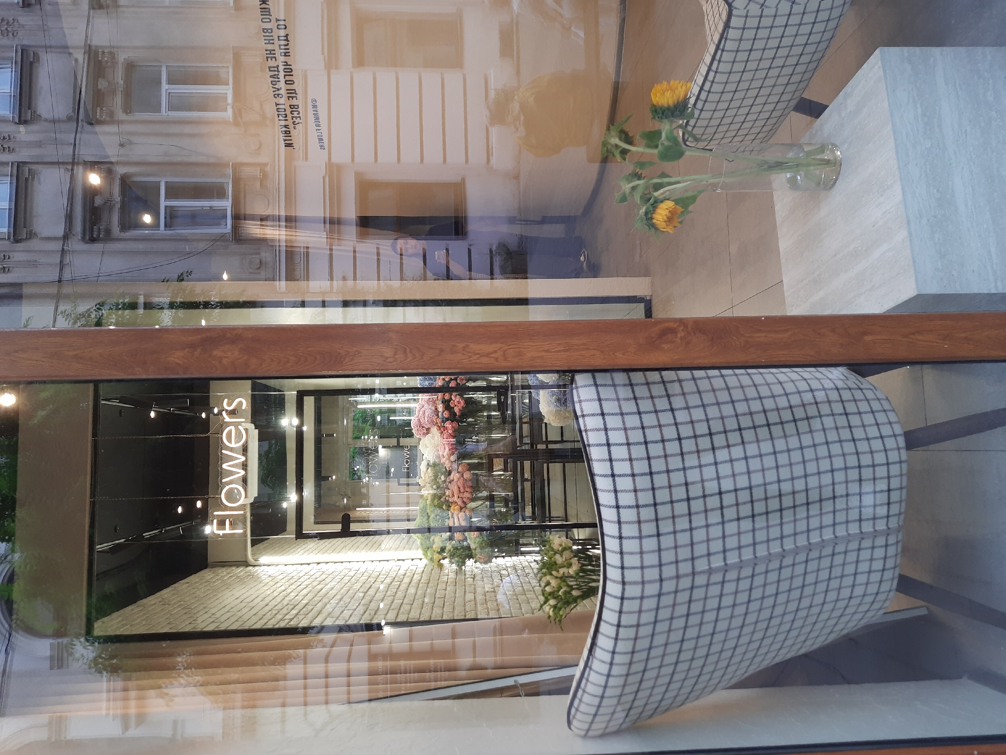 The image shows the storefront of a flower shop in Lviv, Ukraine. Through the window, a neon sign that reads "Flowers" is visible. Inside, shelves are filled with various bouquets of flowers, including roses and hydrangeas, displayed in vases. In the foreground, a checkered armchair sits in front of the window. To the right of the armchair, a small side table holds a glass vase with two sunflowers. Reflected in the window are buildings across the street, one with multiple windows and ornate detailing, and a person standing inside a building holding a smartphone. Text is reflected on the building across the street.
