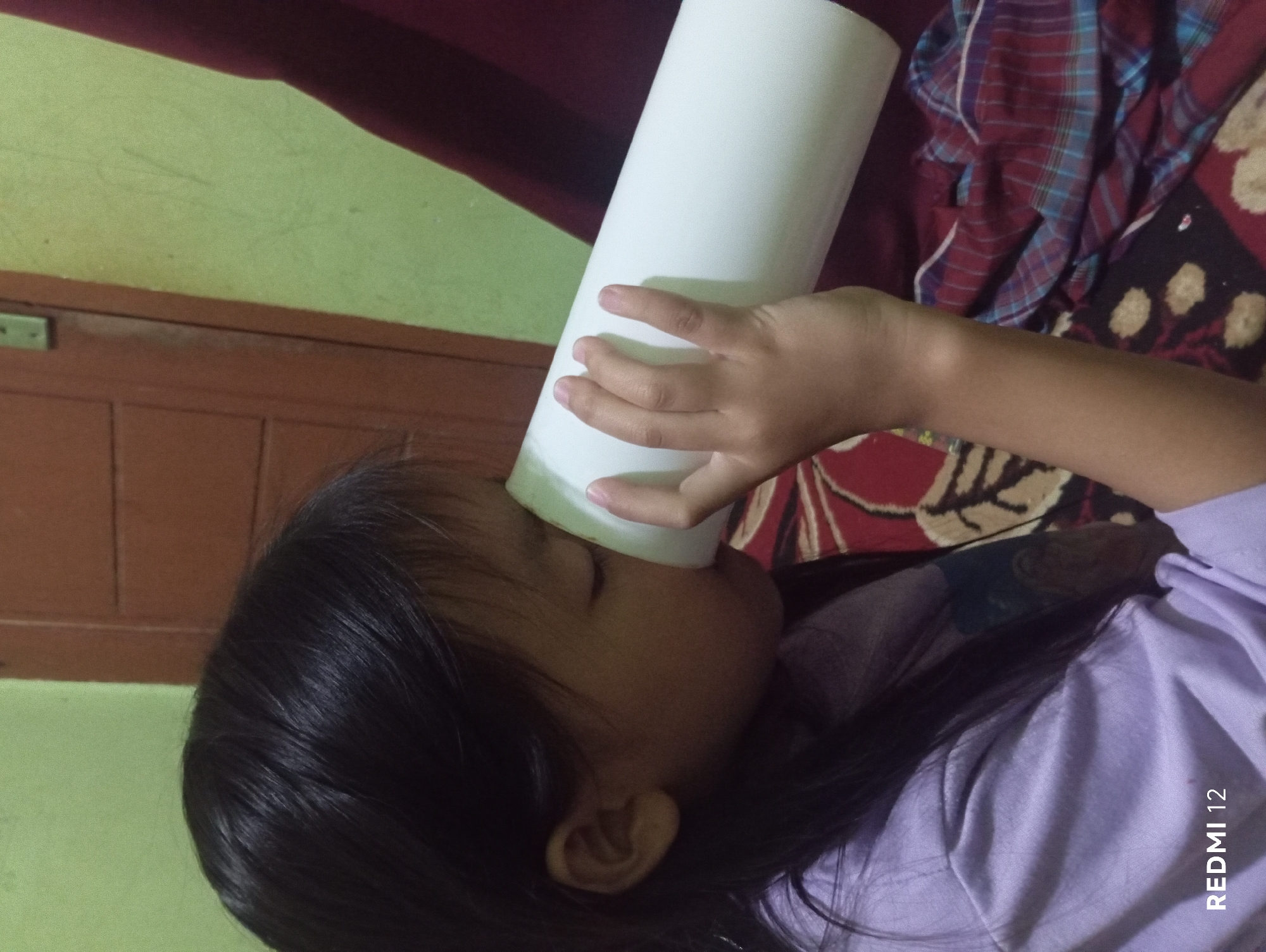 A young girl with dark hair, wearing a light purple t-shirt, is holding a white cylindrical object to her face with her right hand. Her eyes are closed, and she appears to be either drinking from or smelling the object. She is sitting in a room with a green wall and a brown wooden door in the background. A patterned blanket is visible in the foreground. The lower left corner of the image displays the text "REDMI 12". The scene suggests an indoor setting, possibly a home in Pagar Alam, Indonesia, given the device information.