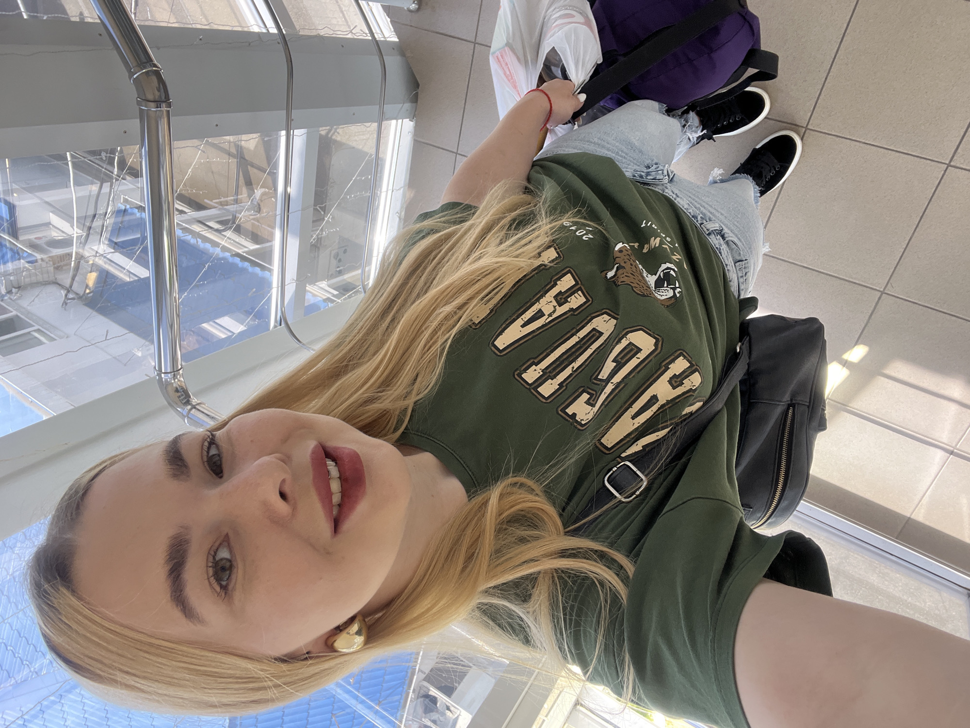 The image shows a young woman taking a selfie in an indoor setting, possibly an airport or a large public building. She has blonde hair and is wearing a green t-shirt with a graphic print of a roaring animal and text that appears to say "NEW YORK". She is also wearing distressed denim jeans and black sneakers. A black backpack is slung over her shoulder, and a purple bag is visible behind her. Her facial expression suggests she is happy and relaxed. The floor is tiled, and there are large windows in the background showing an outdoor area with some industrial structures. The lighting suggests it is daytime. A red string bracelet is visible on her wrist. The setting appears to be modern and clean, with metal railings and a glass enclosure. The context provided is Krasnodar, Russia, which is consistent with the architectural style and the general appearance of the scene.
