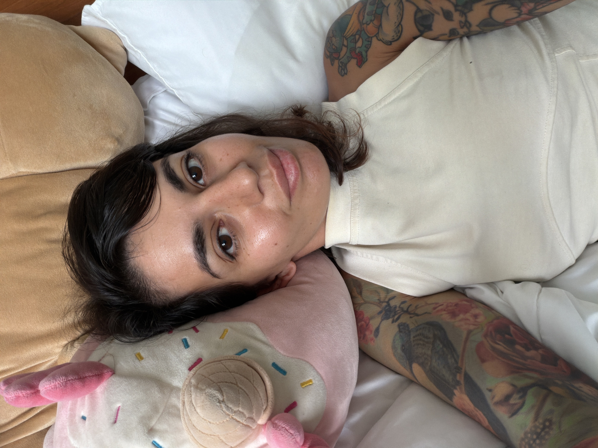 A young adult, with brown hair, brown eyes, and visible facial freckles, is captured in a close-up, overhead shot in Kuta Selatan, Indonesia. The individual is lying on their back, looking directly at the camera with a slight smile. They wear a cream-colored, short-sleeved top. Both arms are heavily tattooed: the right arm (viewer's left) features a green cartoonish creature, while the left arm (viewer's right) displays intricate floral patterns, including red roses and a bird. Their head rests on a plush, pink and white donut-shaped pillow adorned with colorful sprinkles and a light brown concha design. White bed linens and a tan-colored pillow are visible in the background, indicating an indoor, bedroom setting.