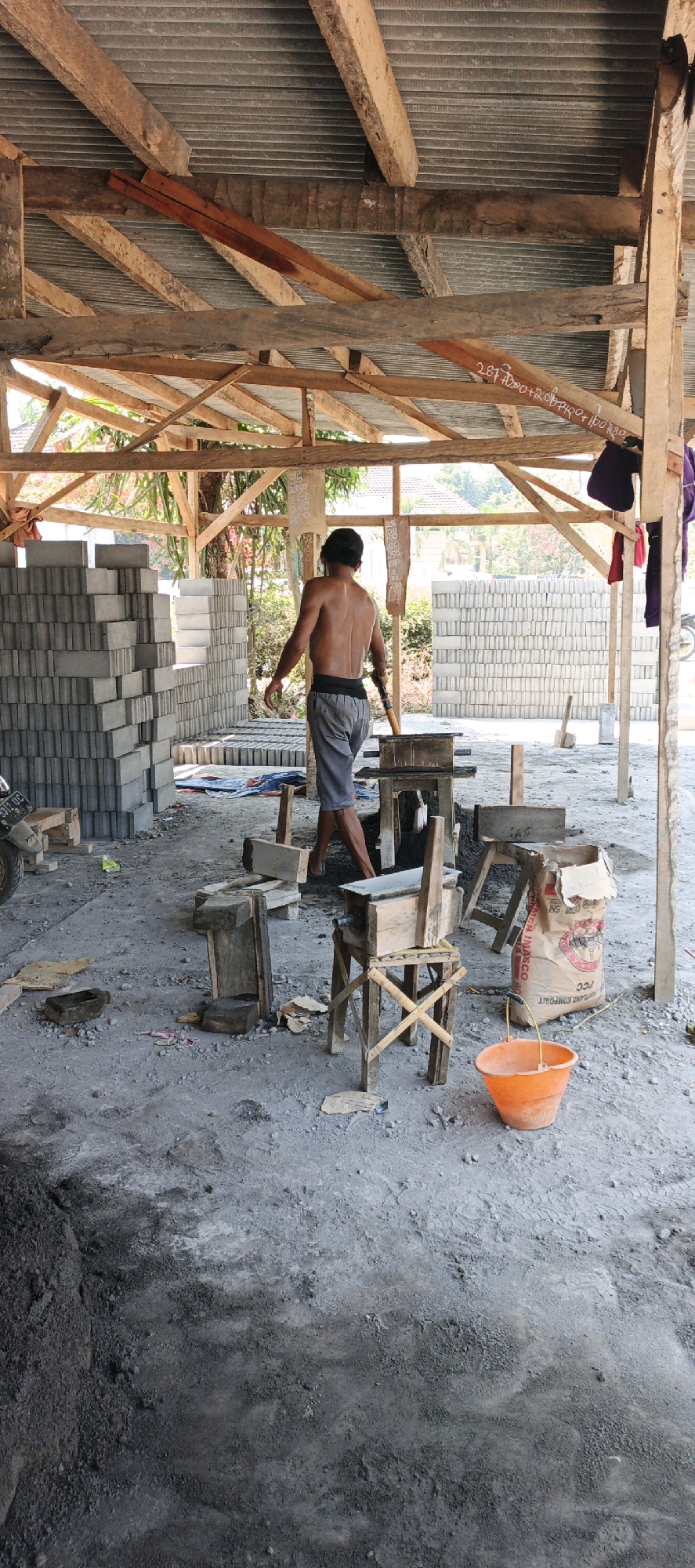 The image depicts a construction site in Kasiyan, Indonesia, possibly a small brick-making facility. A shirtless man wearing grey shorts is walking away from the camera, holding a tool. The environment is an open-air structure with a corrugated metal roof supported by wooden beams. Stacks of grey bricks are visible on both sides of the frame, with more in the background. In the foreground, there are various wooden contraptions that appear to be part of the brick-making process, along with a bag of cement and an orange bucket. The ground is covered in dirt and concrete dust. The scene suggests ongoing construction or manufacturing activity.